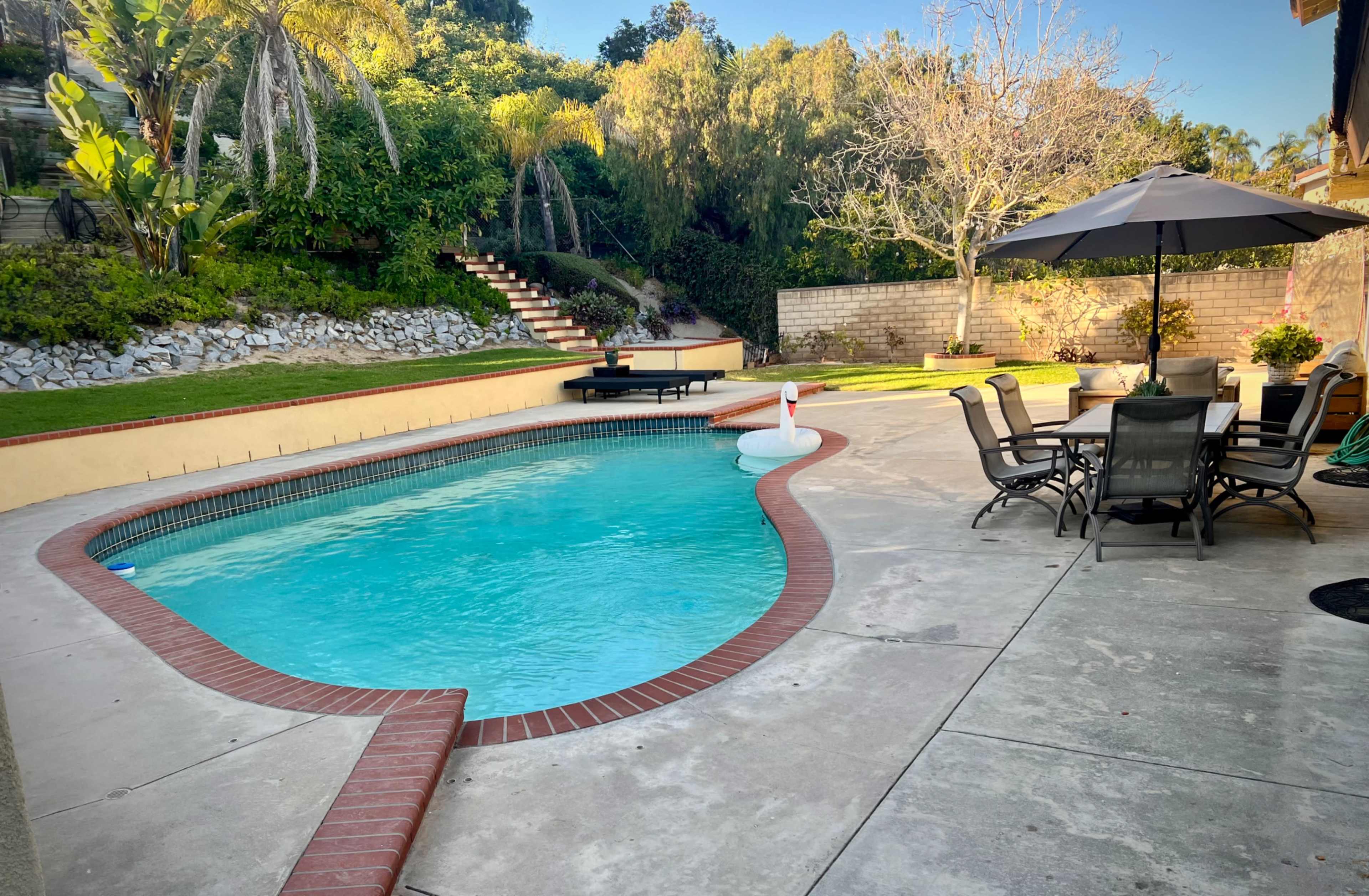 A swimming pool with a brick border is surrounded by a patio and lounge area, featuring an umbrella table and landscaped greenery in the background.