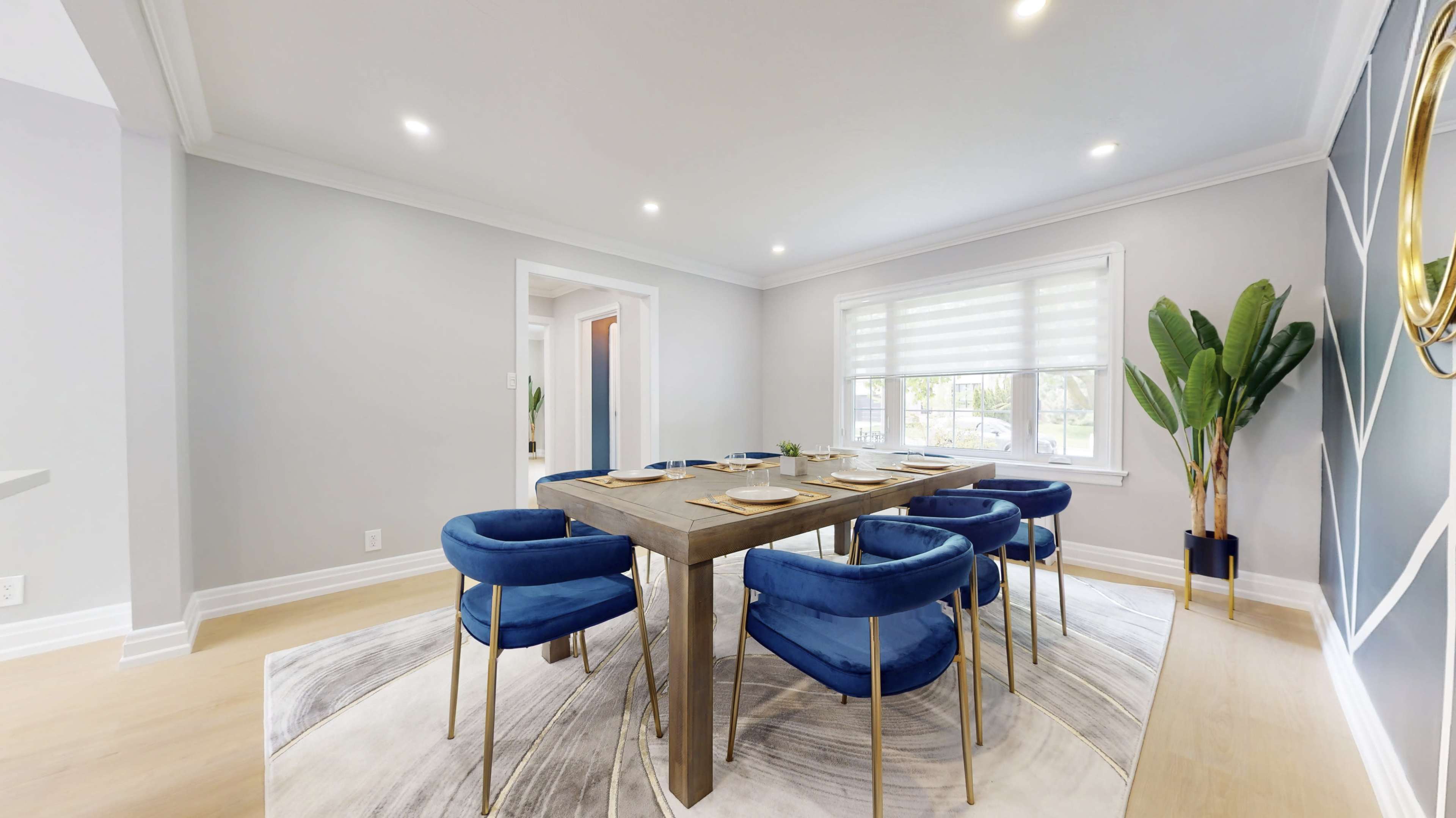 A dining room features a rectangular wooden table surrounded by six blue chairs, set on a textured rug, with a large window and potted plant in the corner.