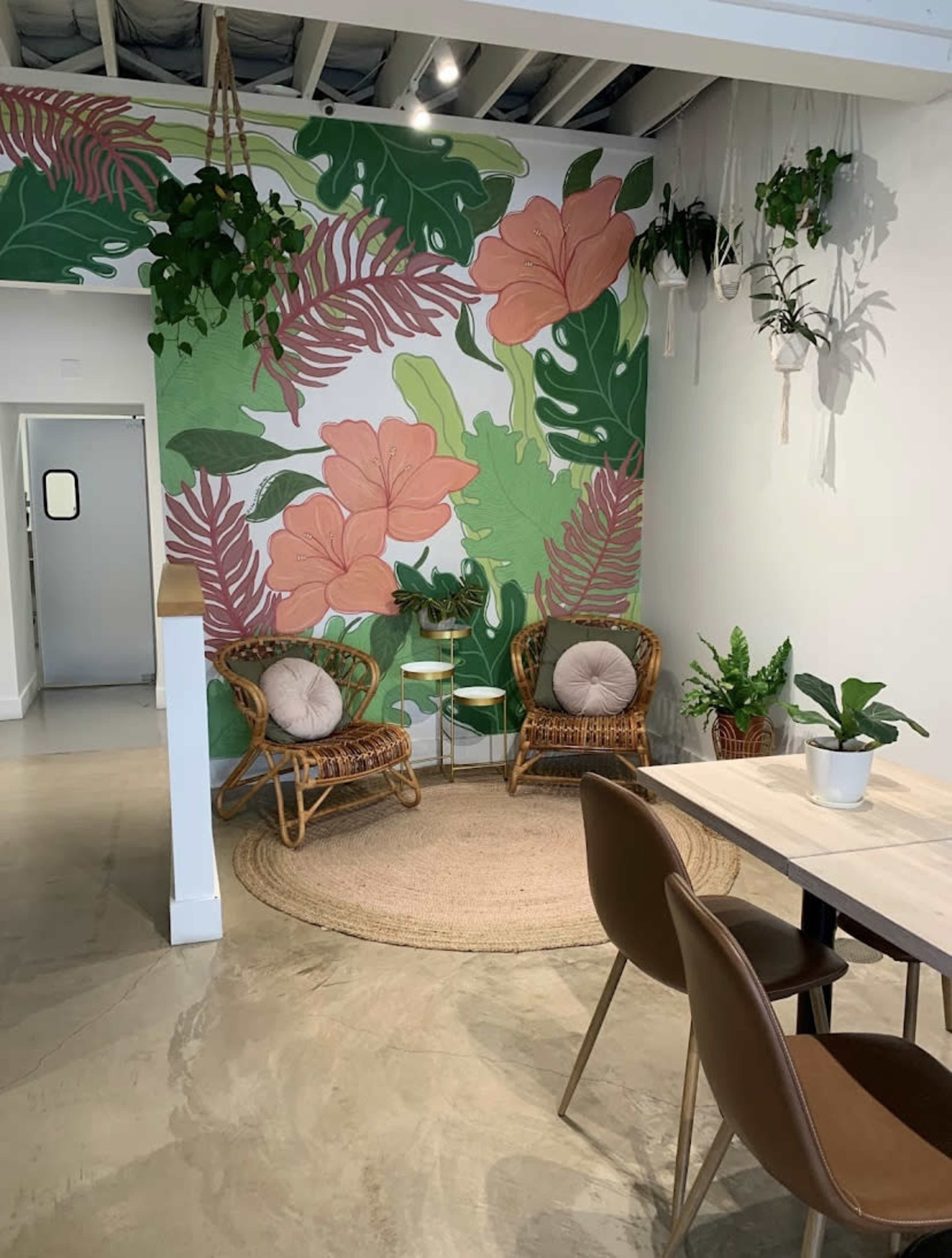 The image shows a cozy corner of a cafe featuring a tropical-themed mural, two rattan chairs, a small table, and various plants in pots.