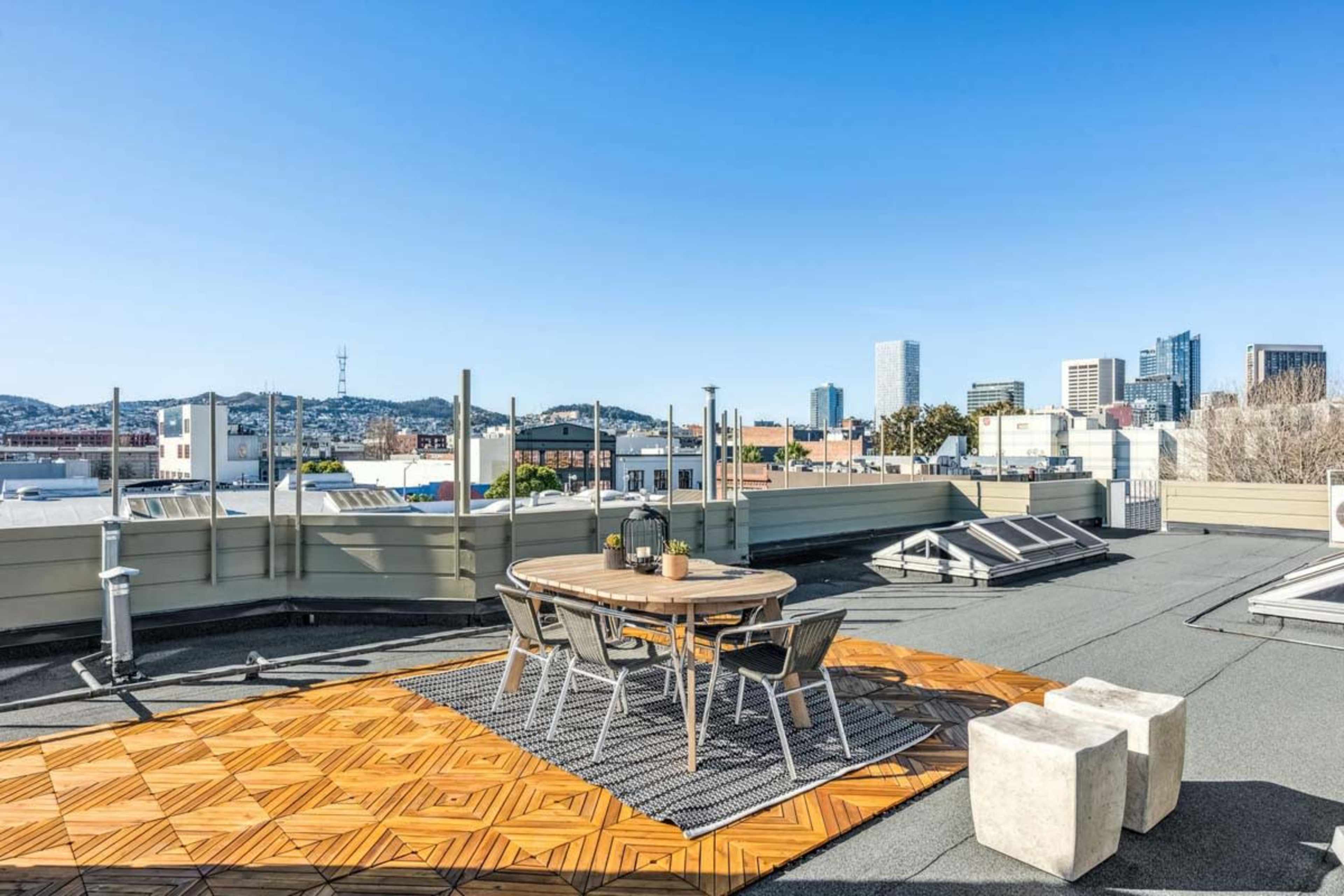 A rooftop terrace features a circular table surrounded by chairs on a wooden platform, with a city skyline visible in the background.