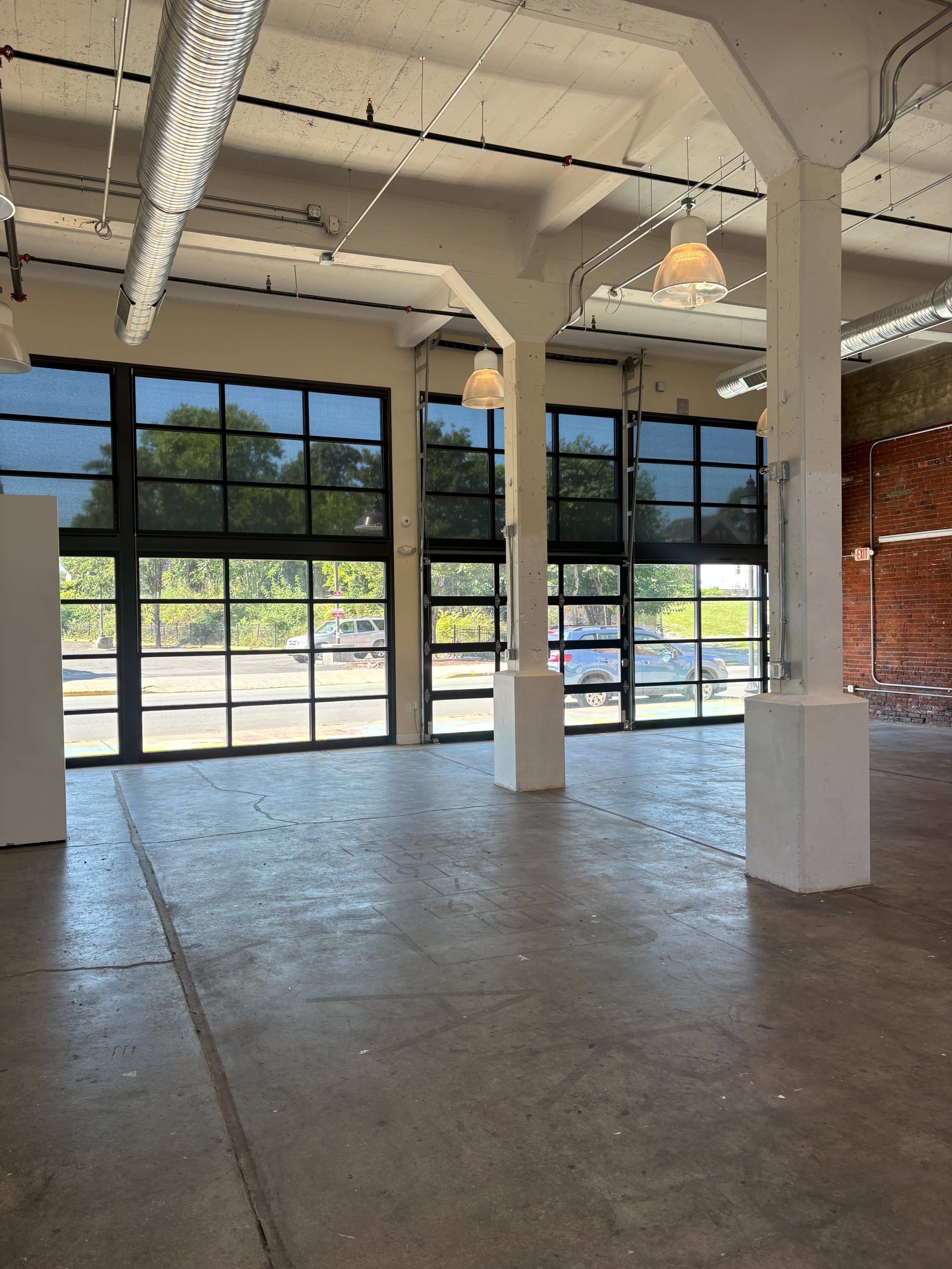 The image shows an empty industrial space with large windows and concrete floors, featuring exposed beams and a brick wall.