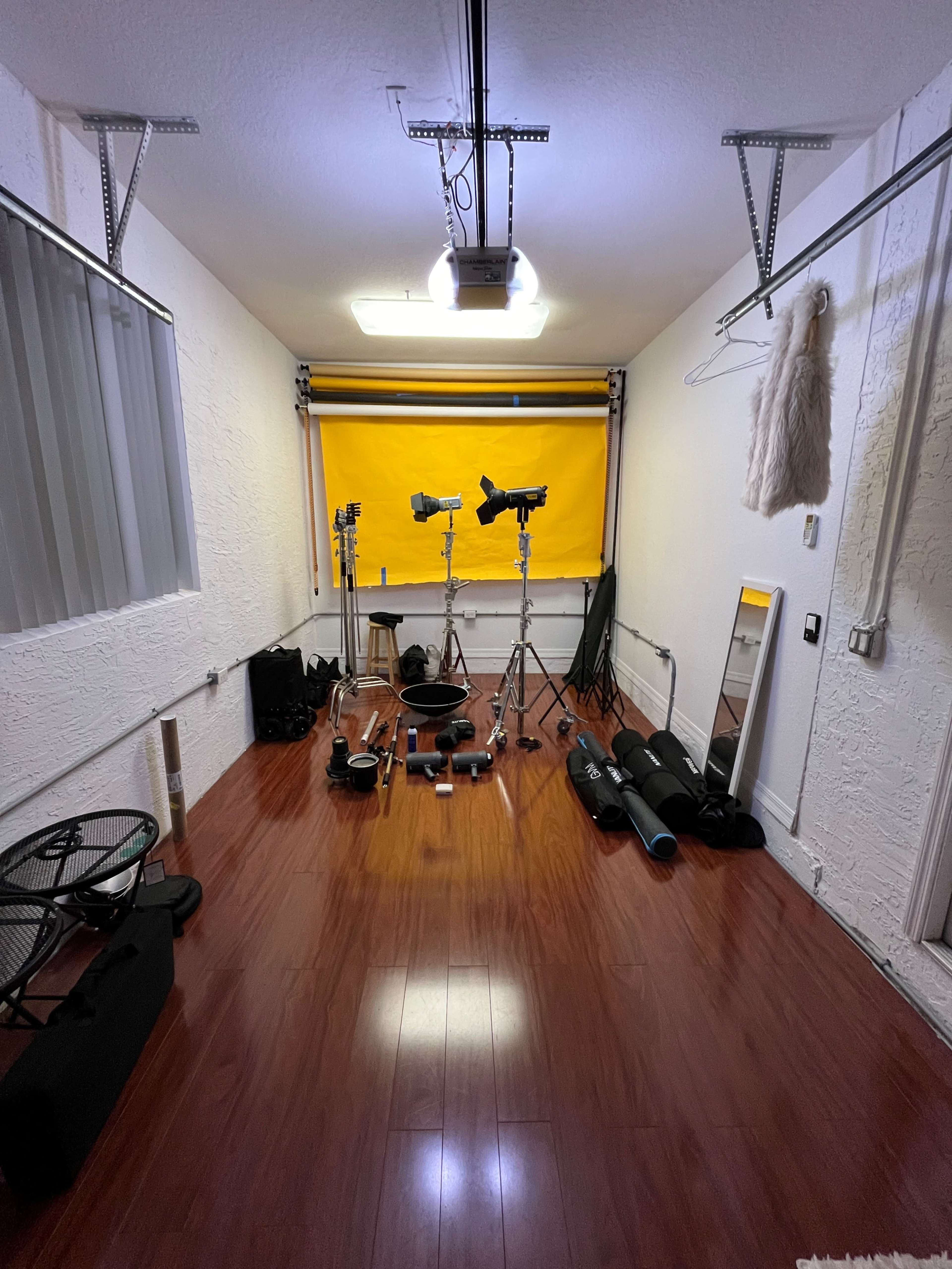 A studio space with three light stands, various photography equipment on the floor, a large yellow backdrop, and a full-length mirror against the wall.