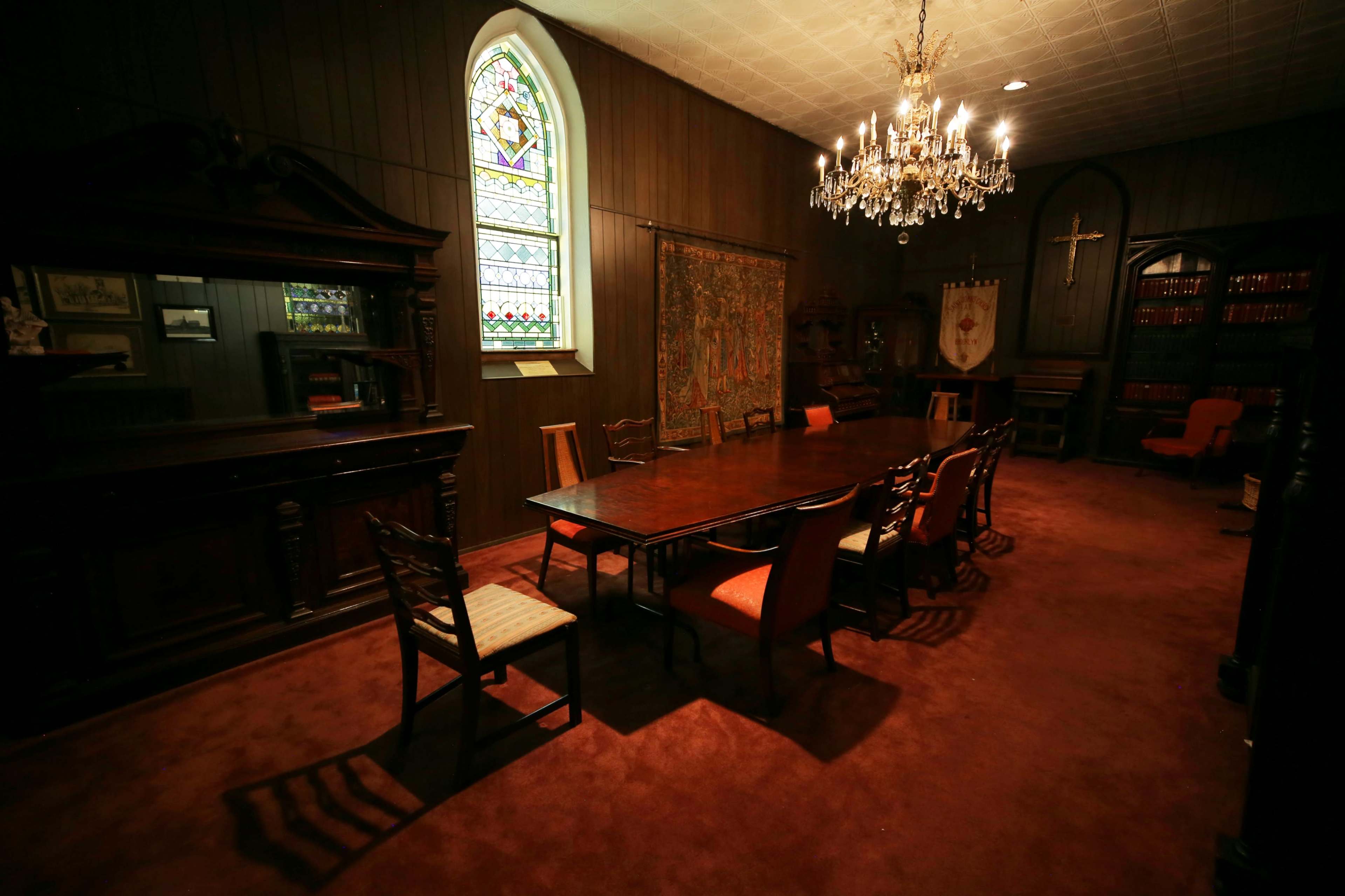 A large, dimly lit meeting room features a long wooden table surrounded by chairs, with a stained glass window and a chandelier overhead.