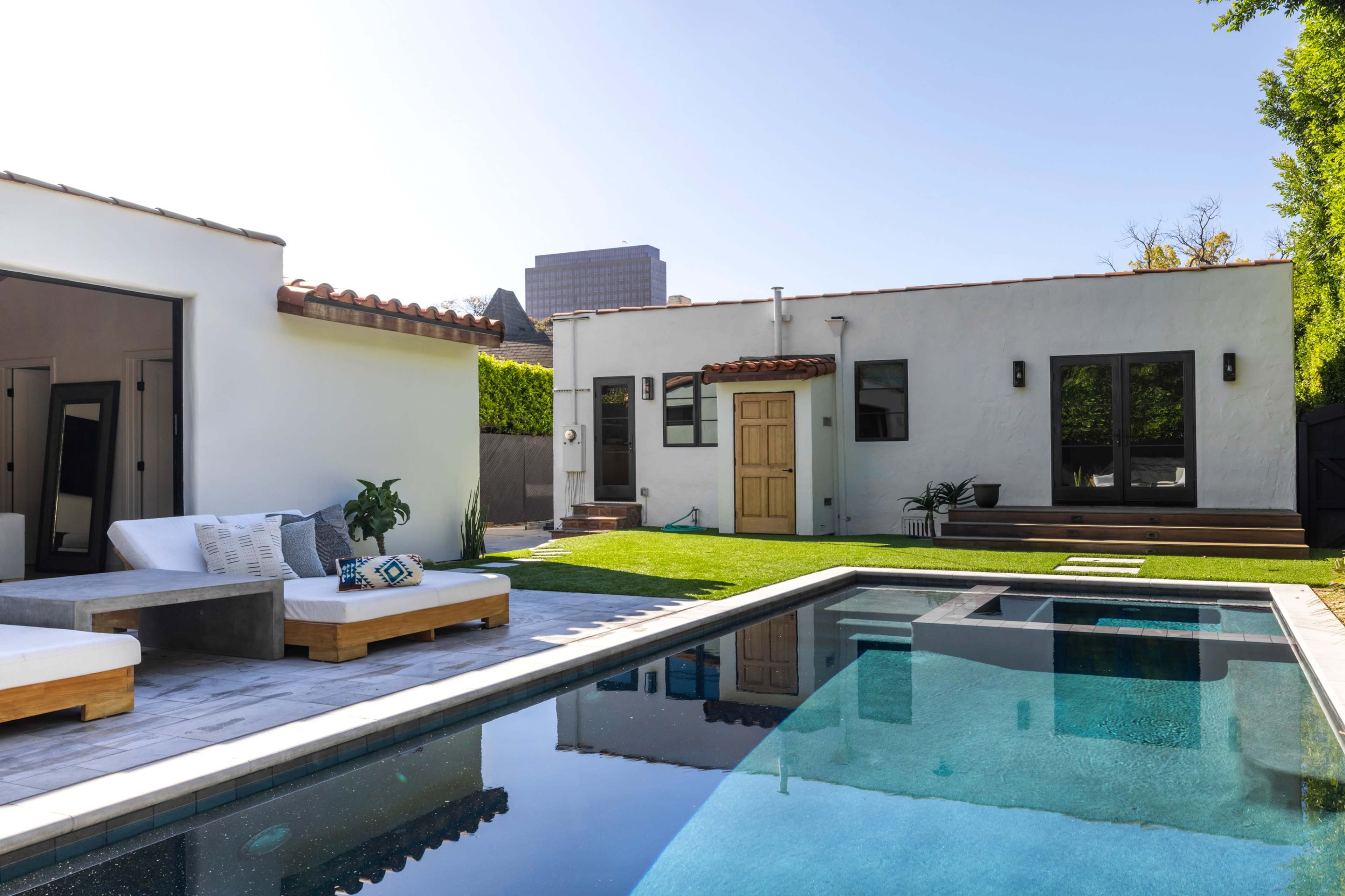 A modern backyard features a swimming pool, a wooden deck, and a white house with a brown door and large windows.