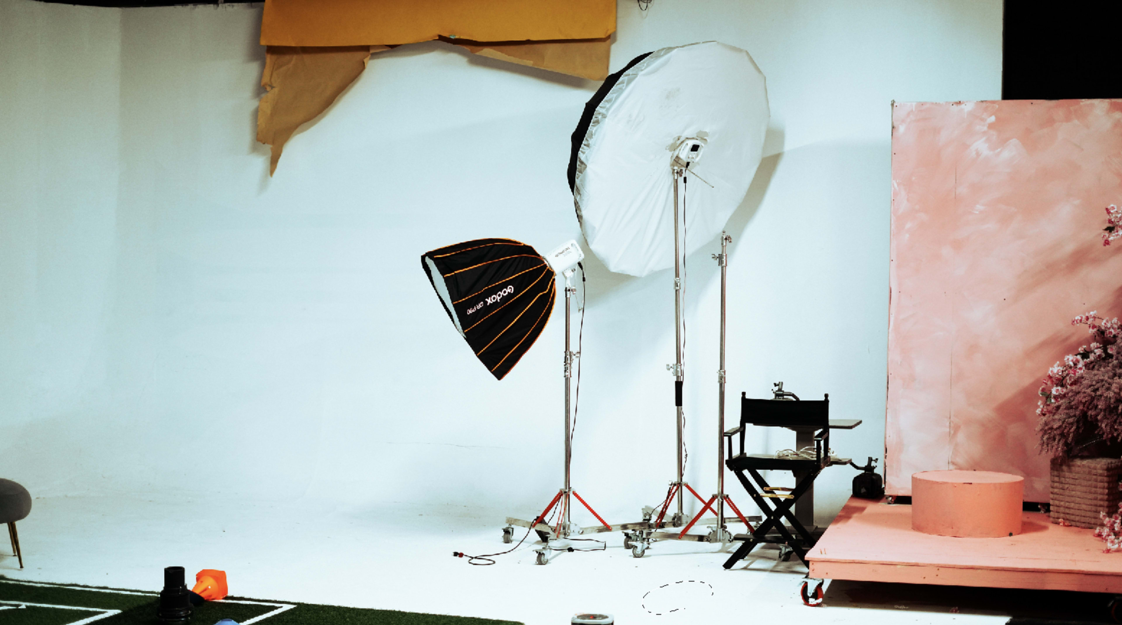 The image shows a photography studio setup with two lighting softboxes, a director's chair, and a pink backdrop.