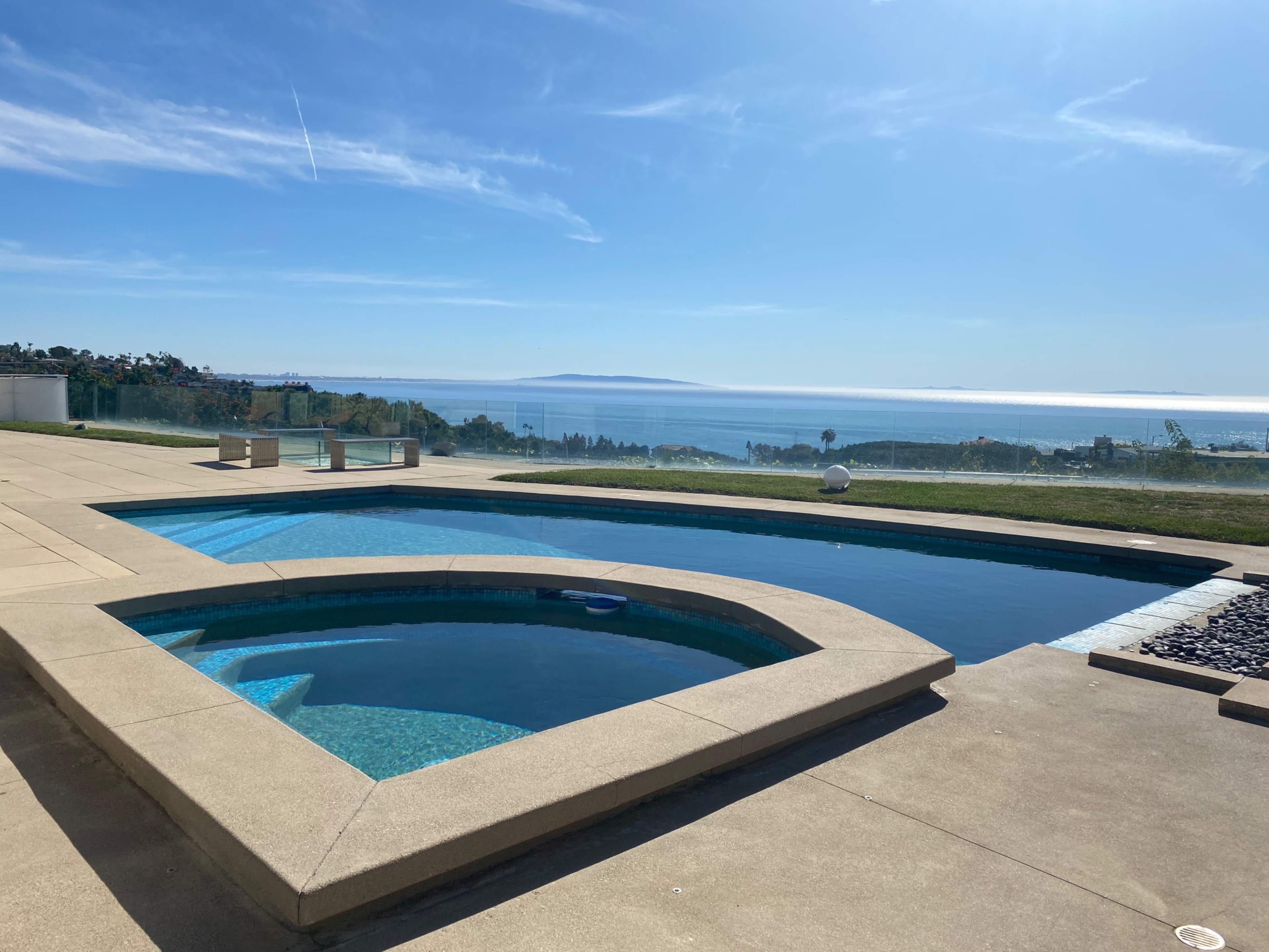 A clear view of a swimming pool and hot tub overlooks a serene ocean landscape under a blue sky.
