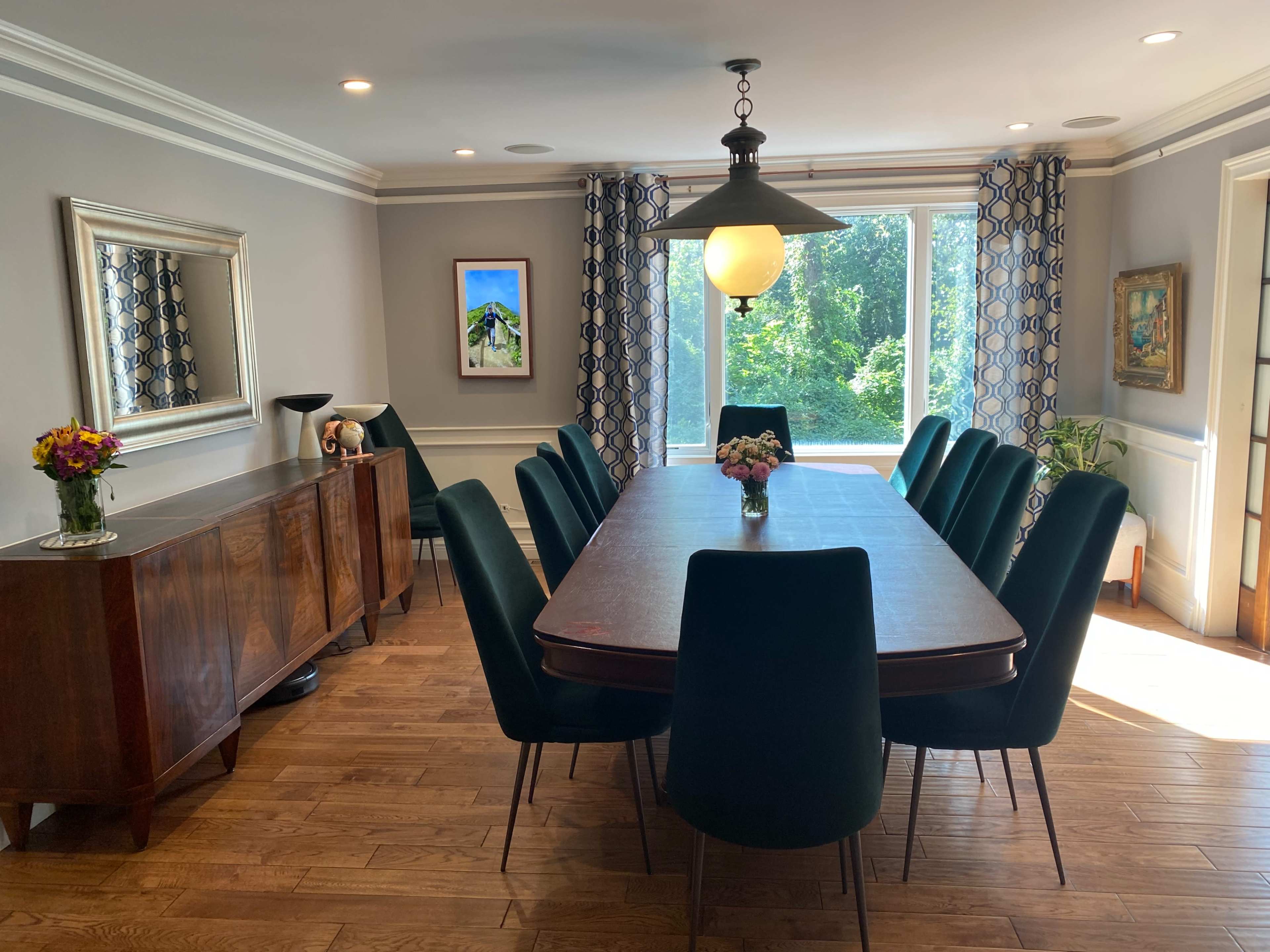 The dining room features a long wooden table surrounded by green upholstered chairs, with a large window overlooking a garden and decorative artwork on the walls.