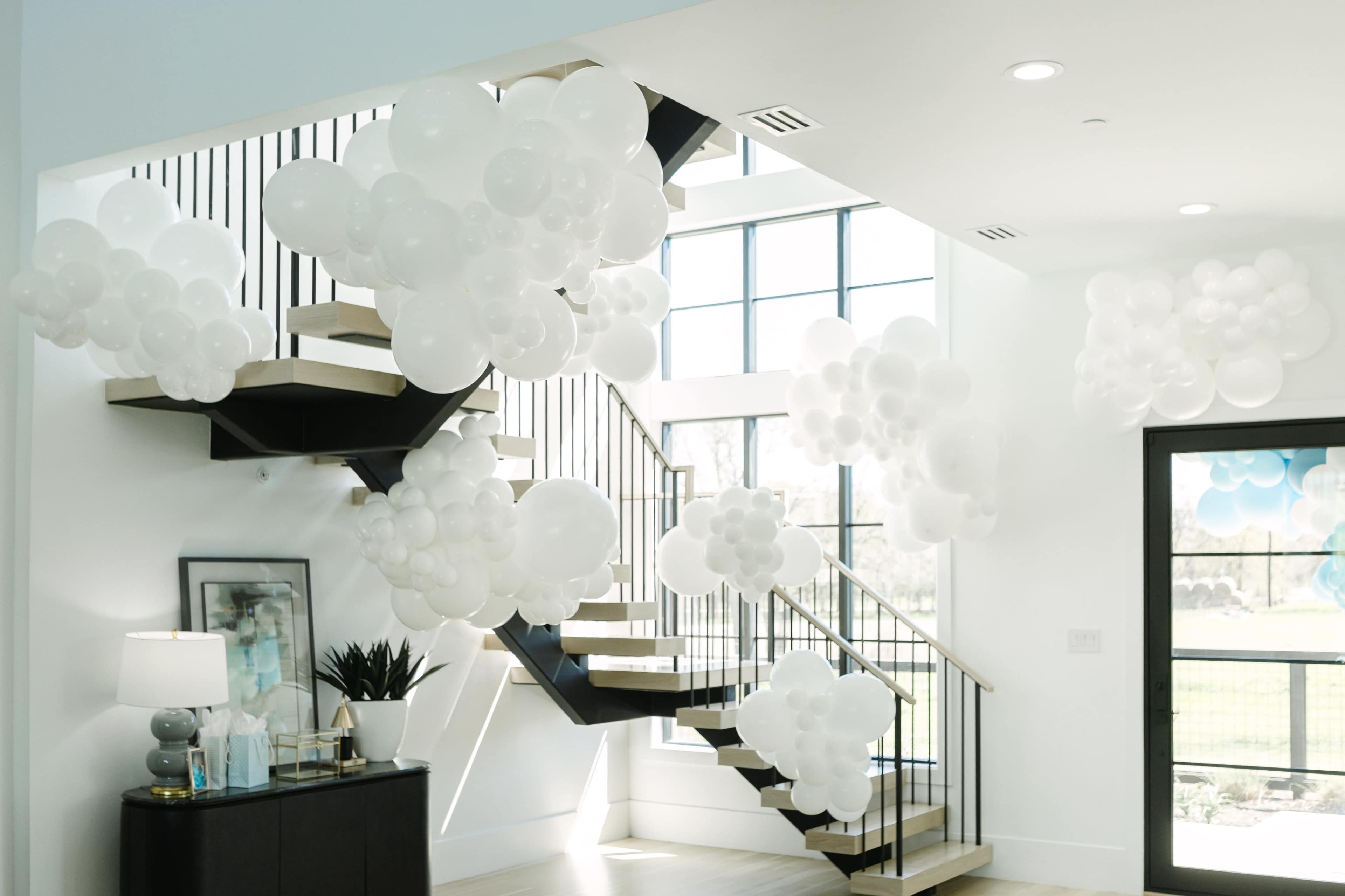 The image shows a modern interior space with a staircase adorned with clusters of white balloons and natural light streaming through large windows.