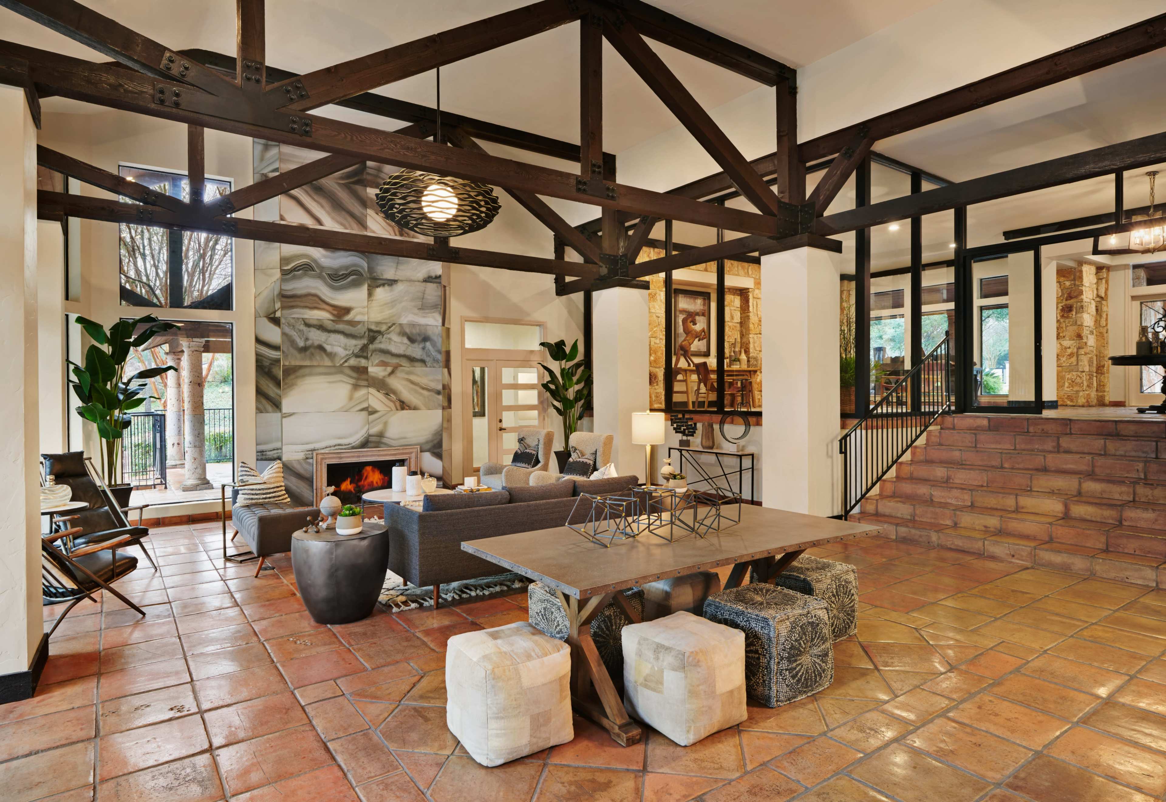A spacious living area with a high ceiling, featuring large windows, a stone fireplace, and a mix of modern furniture, including a large table and comfortable seating.