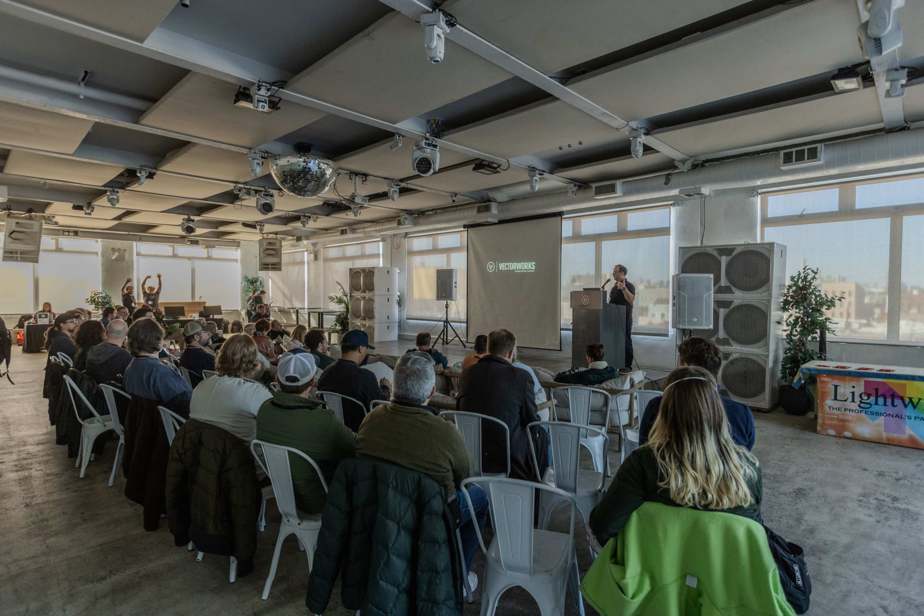 A speaker presents on stage to an audience seated in white chairs in a well-lit conference room.