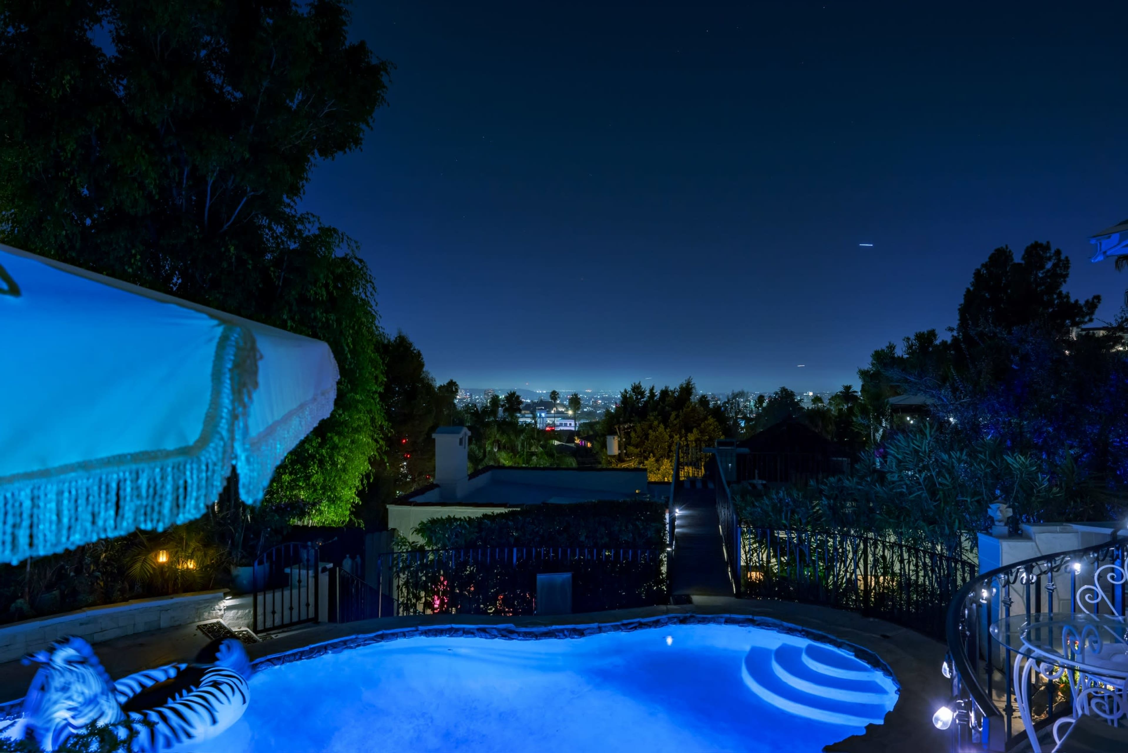 A swimming pool is illuminated at night, with city lights visible in the background and a hillside framed by trees.