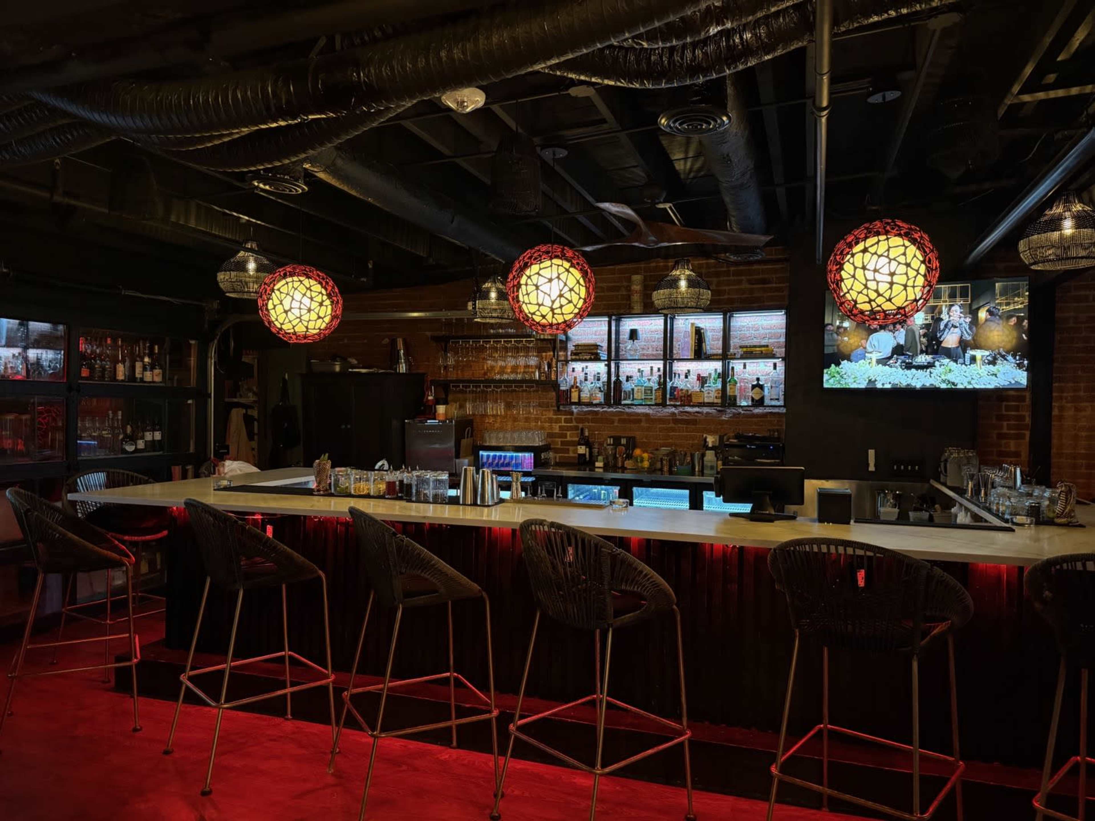 The image shows a dimly lit bar with a long counter featuring high stools, decorative pendant lights, and shelves stocked with various bottles behind it.