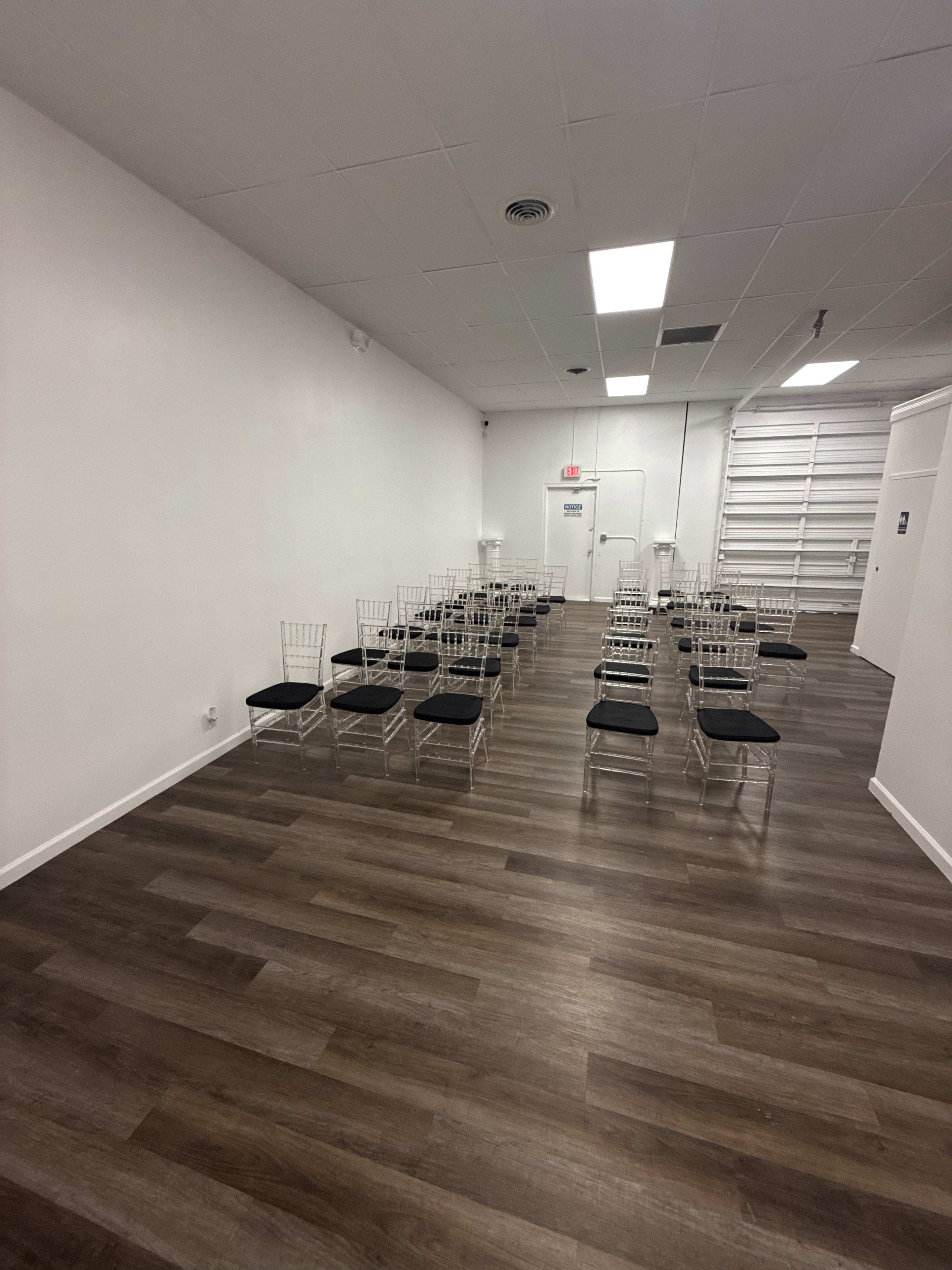 The image shows a narrow room with white walls and a wooden floor, arranged with multiple rows of clear plastic chairs facing the far wall.