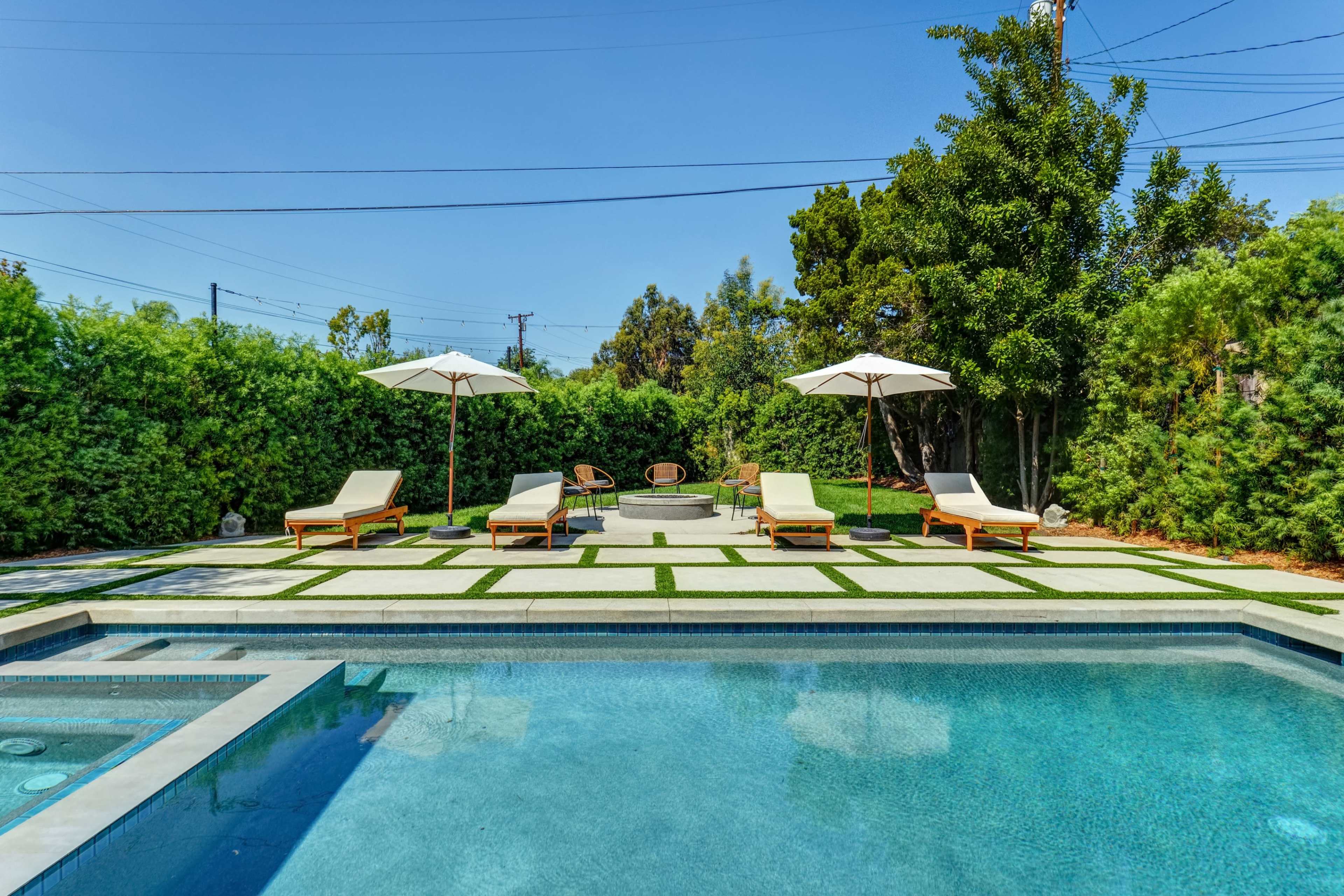 A pool area with four lounge chairs and two umbrellas surrounded by greenery and a patio paved with stone.