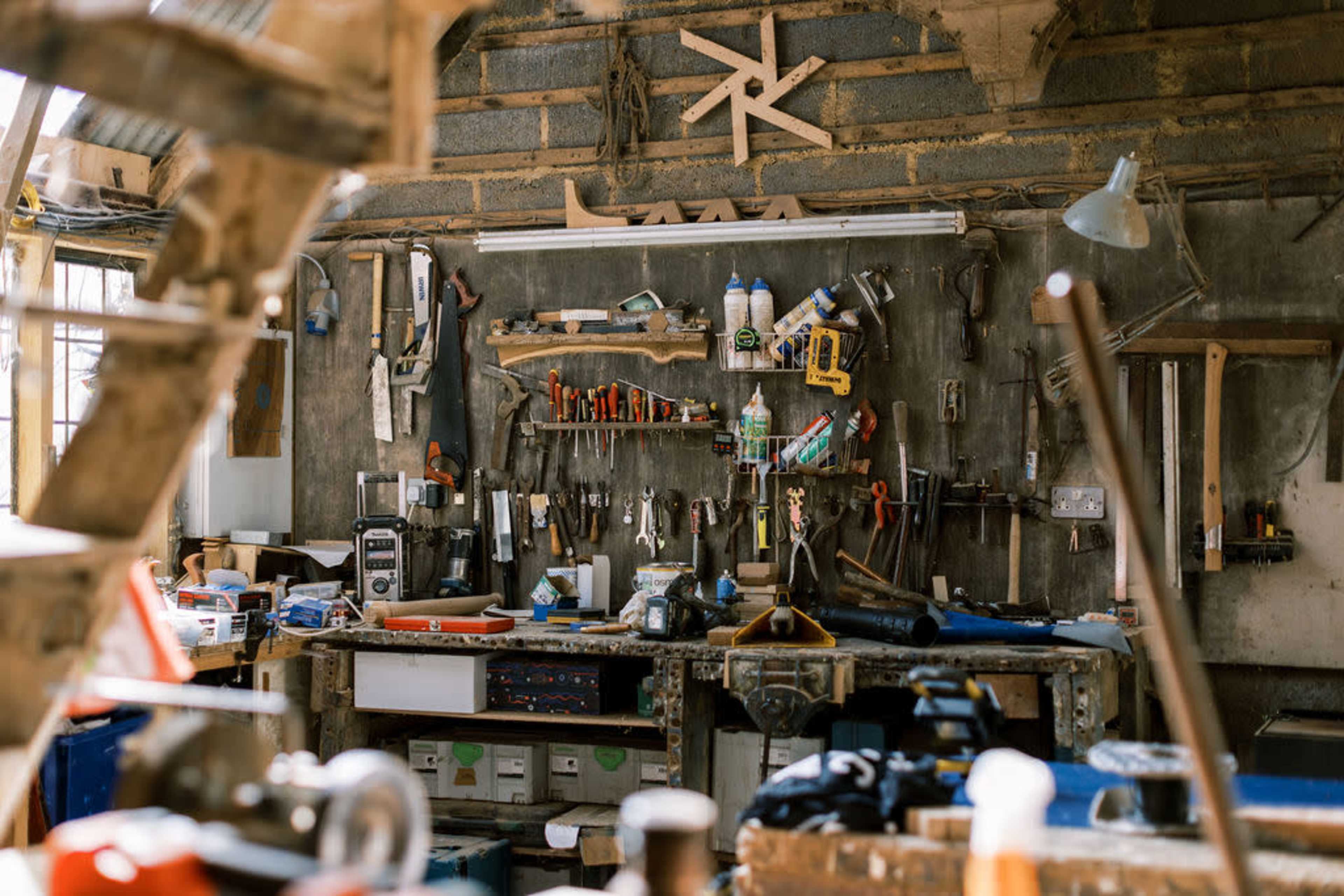 A cluttered workbench is filled with various tools and equipment, with a wall of hand tools organized above it.