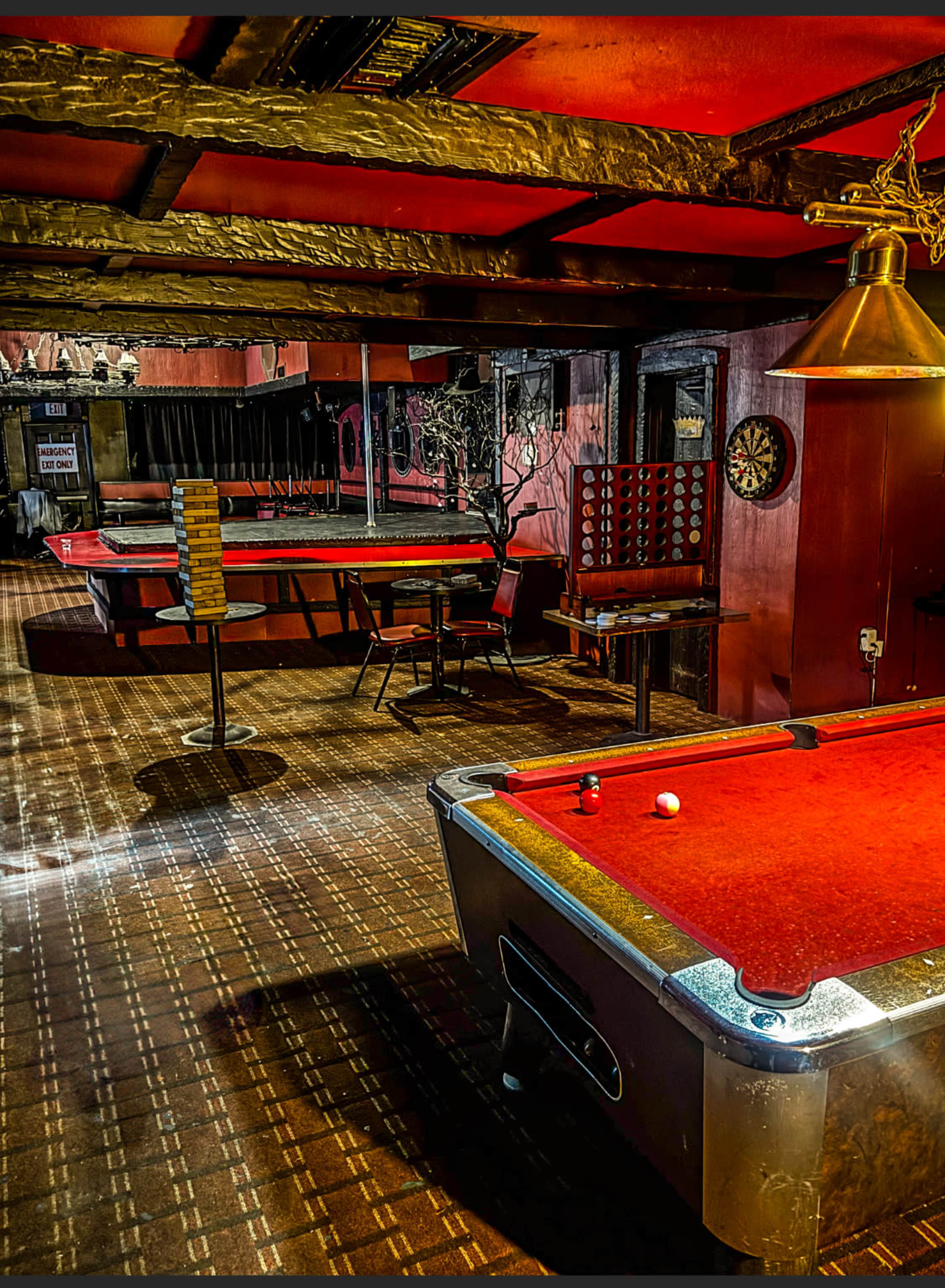 The image depicts an empty pool hall featuring several billiard tables surrounded by chairs and a dartboard on the wall.
