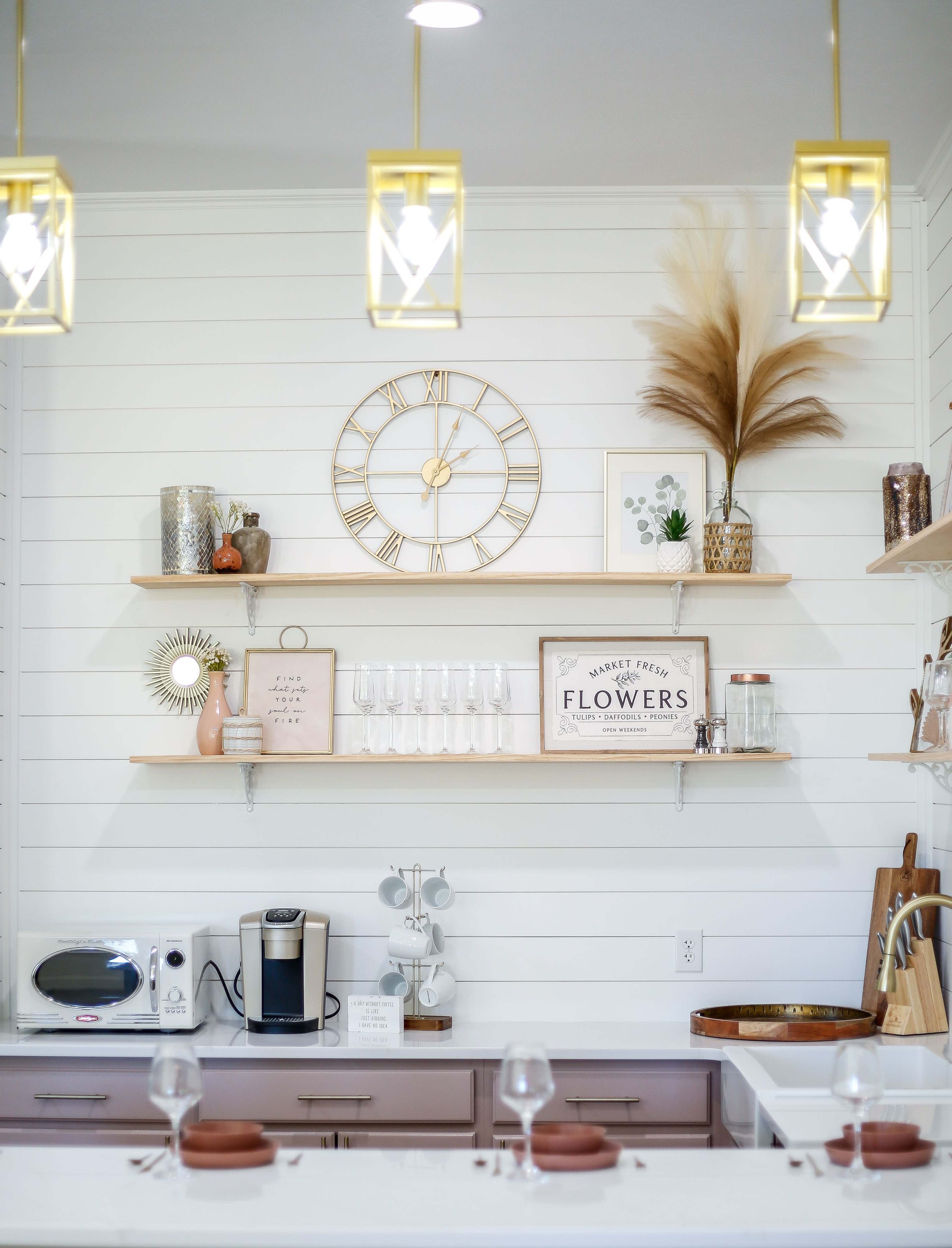 A modern kitchen scene features wooden shelves holding decorative items, a clock, and glassware, with a coffee maker on the countertop.