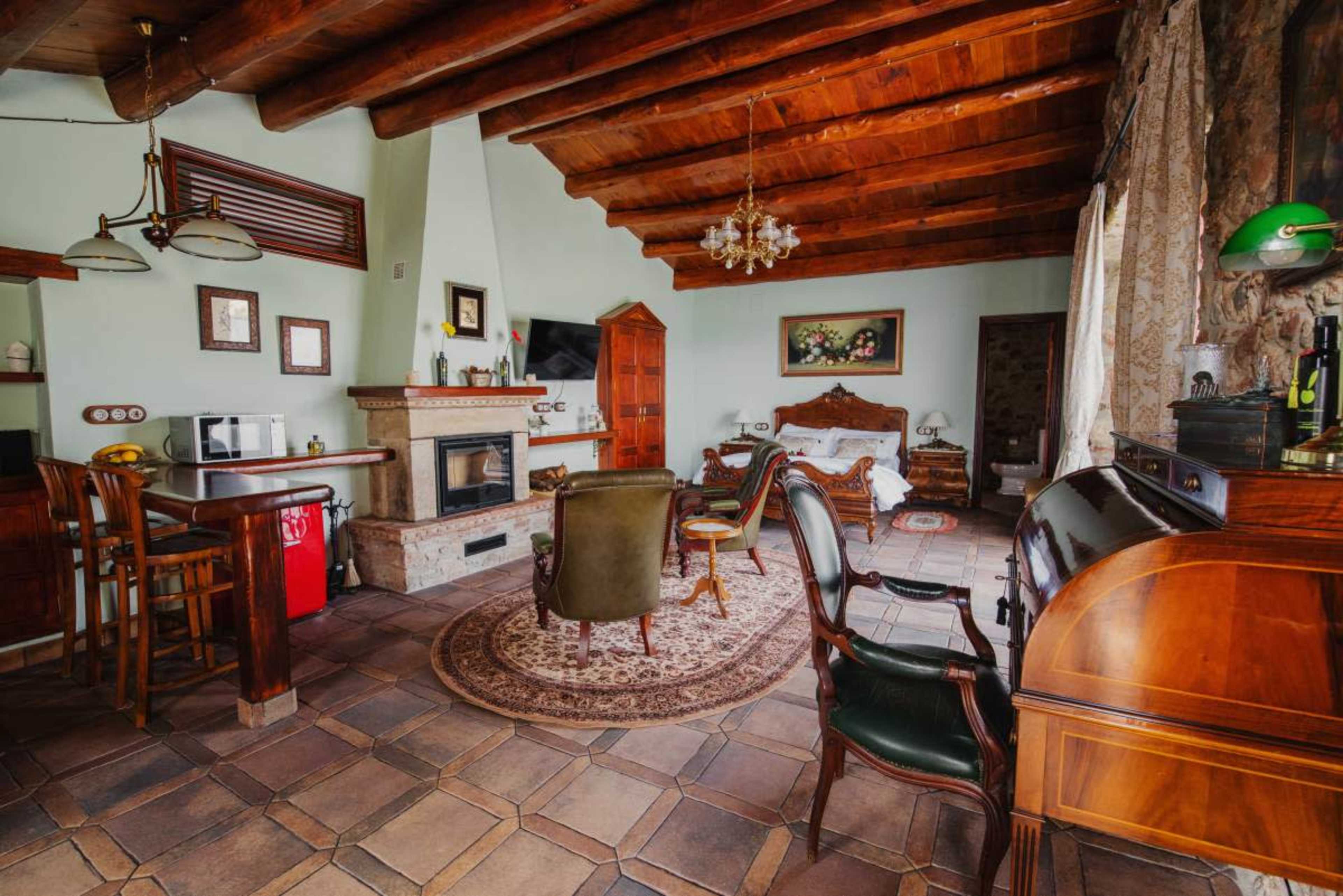 The image shows a cozy living room with wooden beams, a fireplace, and a mix of antique furniture including a bed, chairs, and a small dining table.