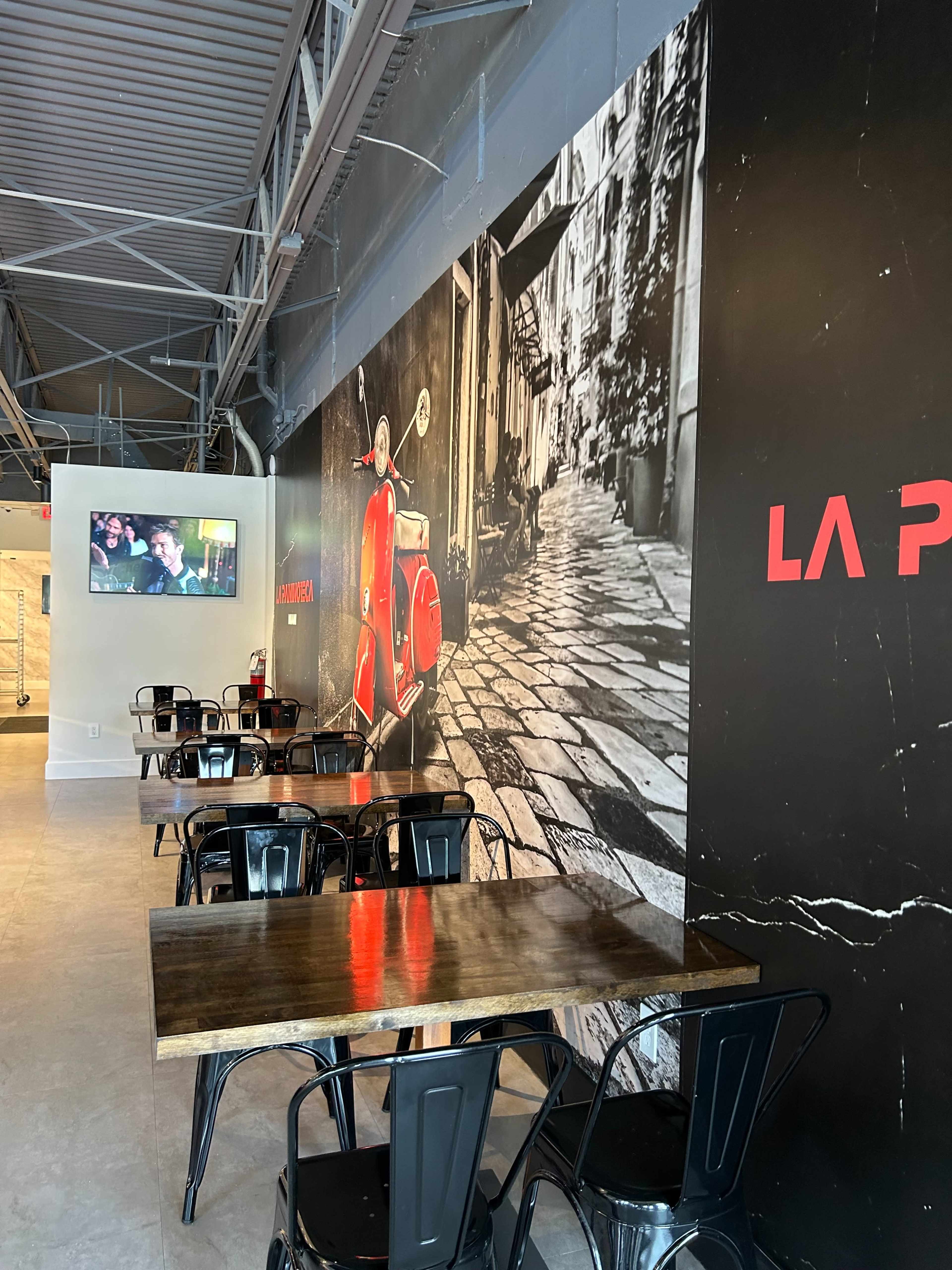 The image shows a modern cafe interior with black metal chairs and wooden tables, featuring a large mural of a red scooter on a cobblestone street.
