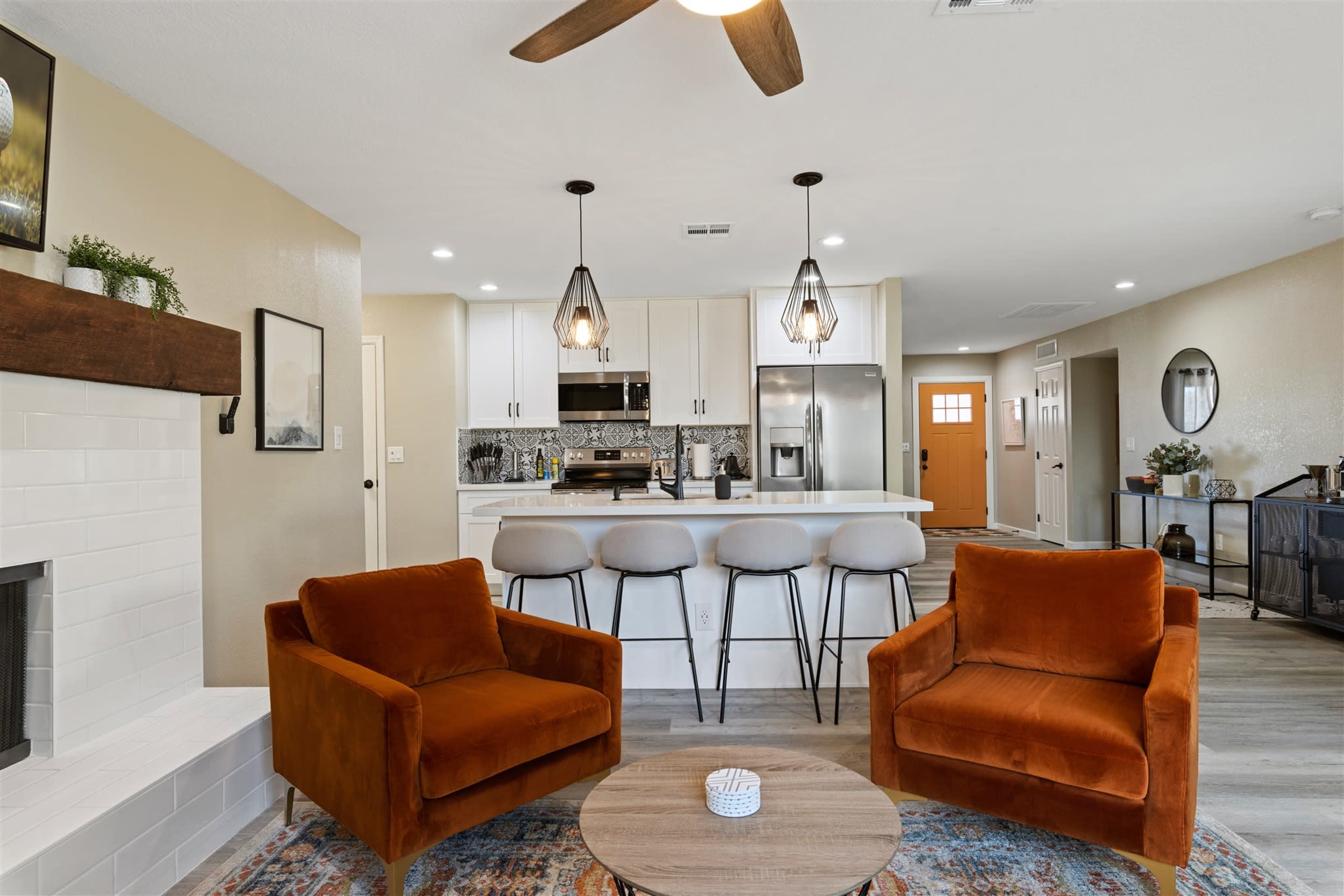 A modern living room featuring two orange armchairs, a round wooden coffee table, and a kitchen area with white cabinetry and stainless steel appliances in the background.