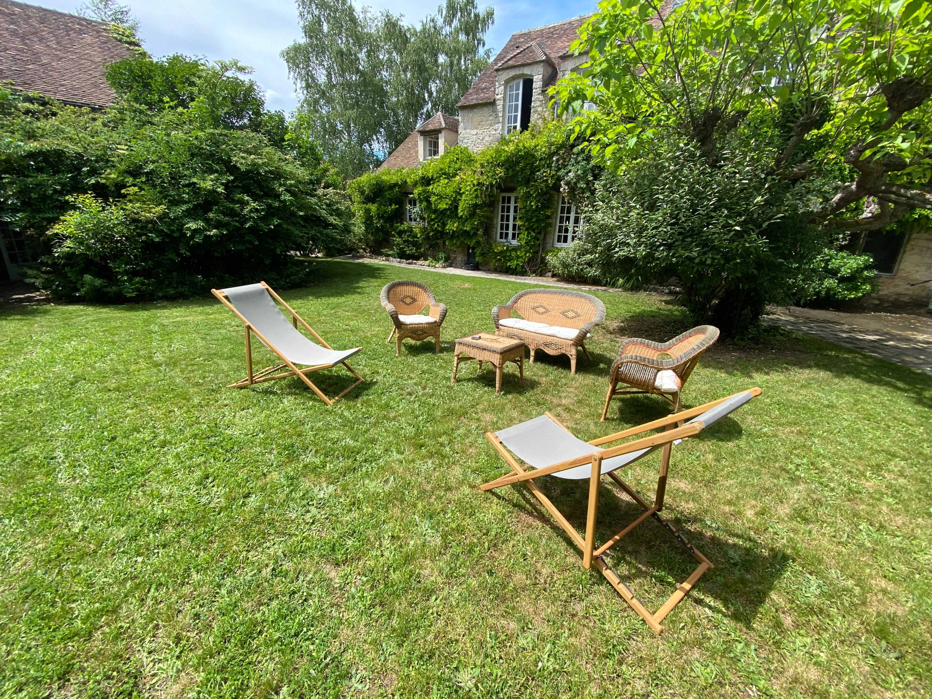 A garden scene featuring several outdoor chairs and loungers arranged on a grassy lawn amidst lush greenery and a stone building in the background.