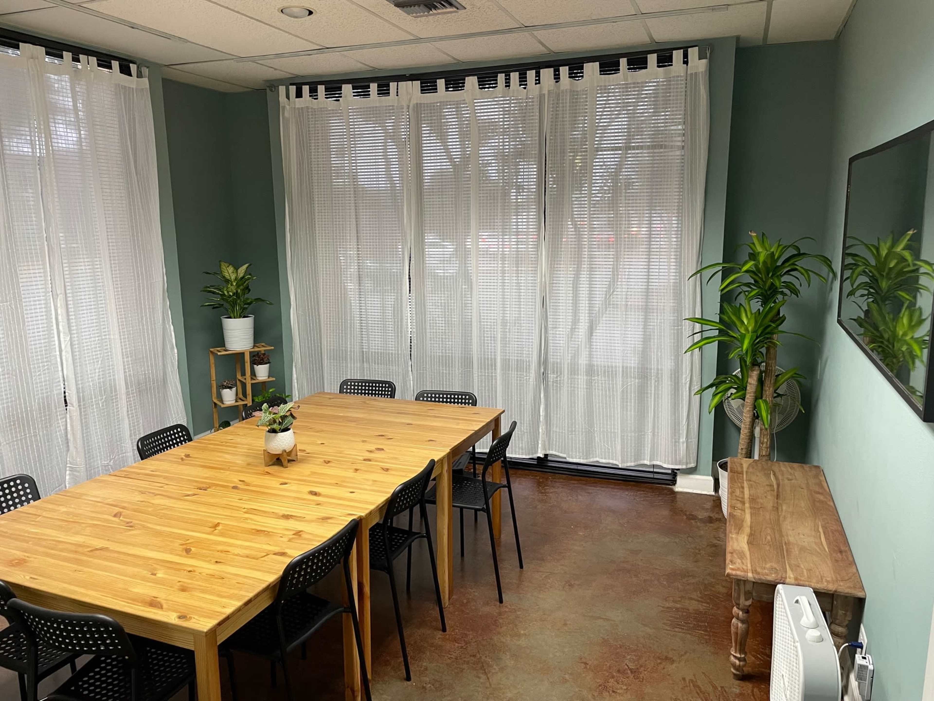 The image shows a conference room with a large wooden table surrounded by black chairs, brightened by natural light from large windows with sheer curtains.