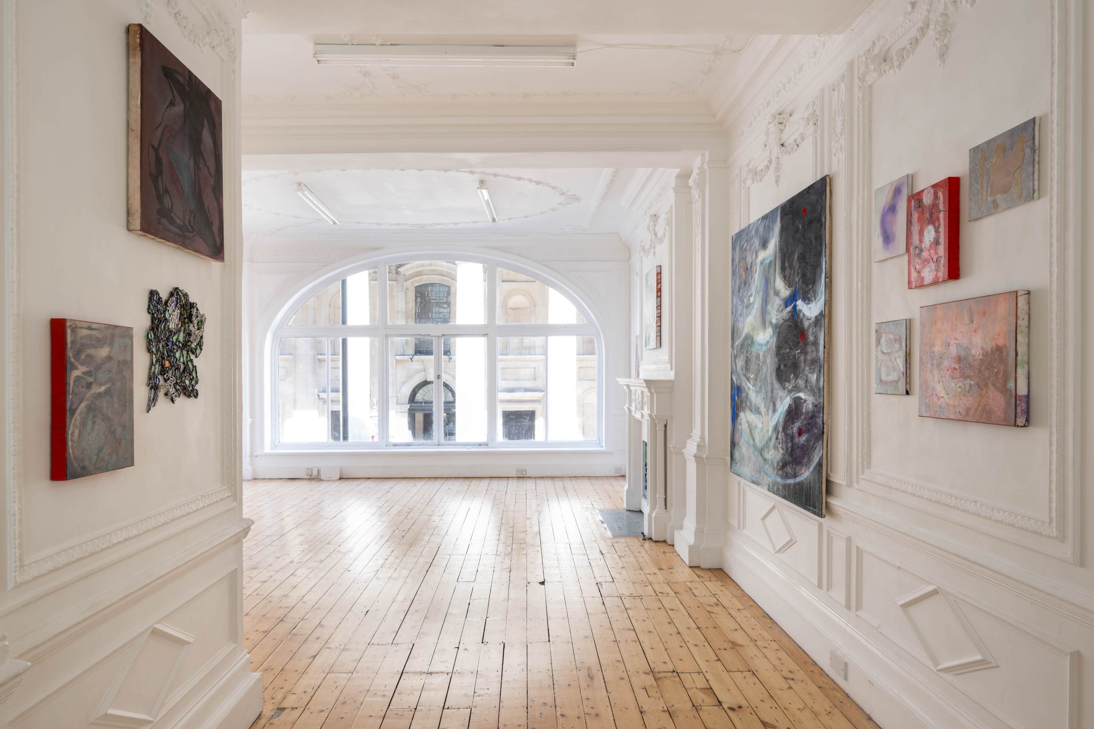 The image shows a bright art gallery with wooden flooring, featuring various paintings hung on the white walls and large windows allowing natural light to flood the space.