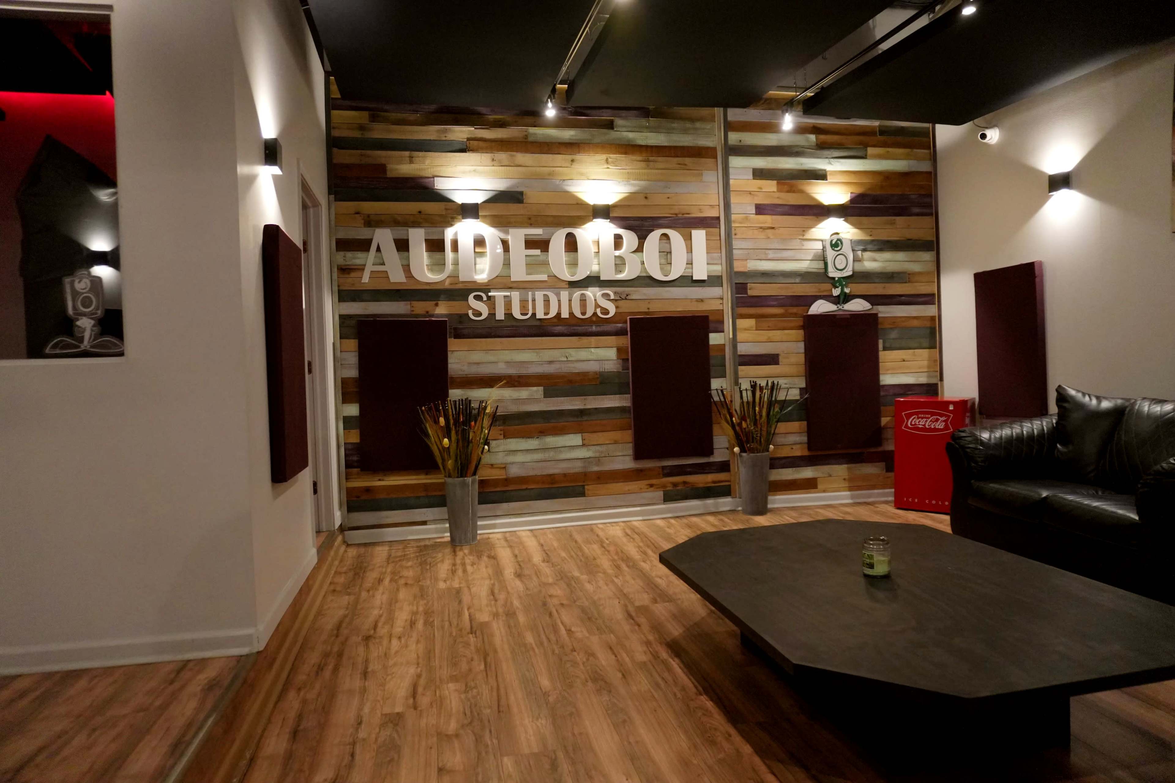 A modern studio interior featuring a wooden accent wall with the text "AUDEOBOI STUDIOS," a black leather couch, a low coffee table, and decorative plants.
