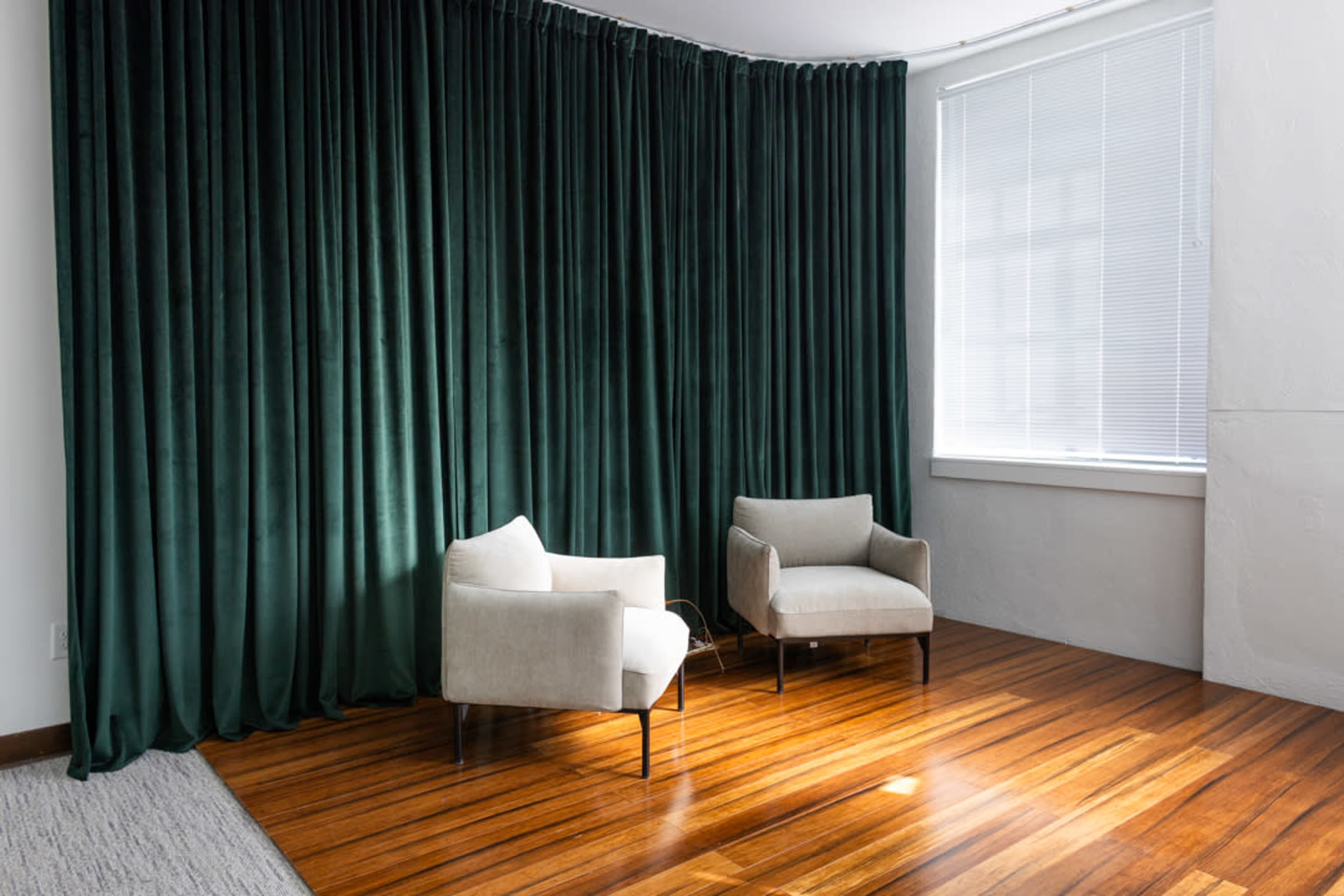 The image shows two light-colored armchairs positioned against a backdrop of deep green curtains in a well-lit room with wooden flooring.