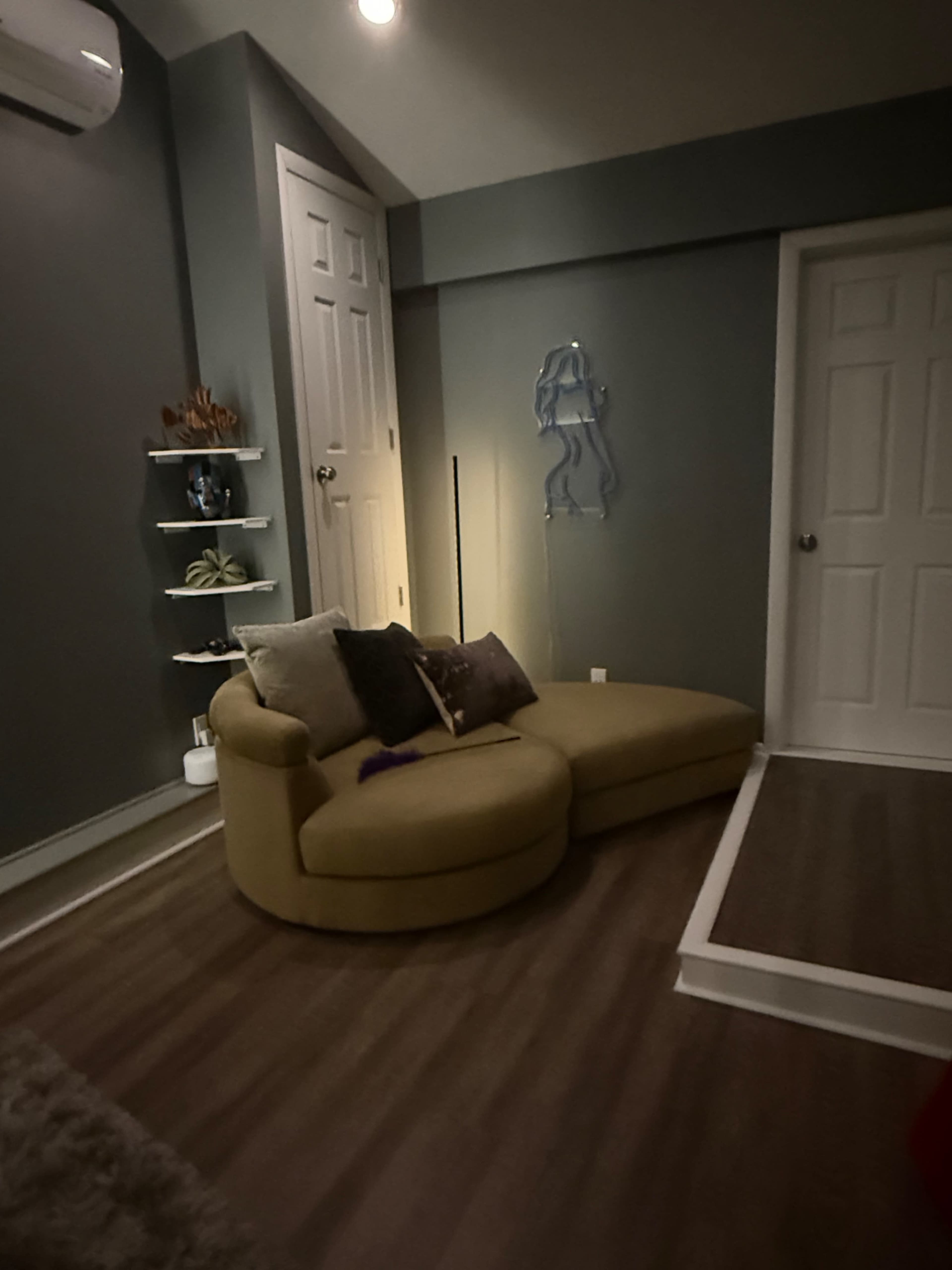 The image shows a cozy corner of a room featuring a curvy yellow couch, a decorative shelf on the wall, and a closed door.