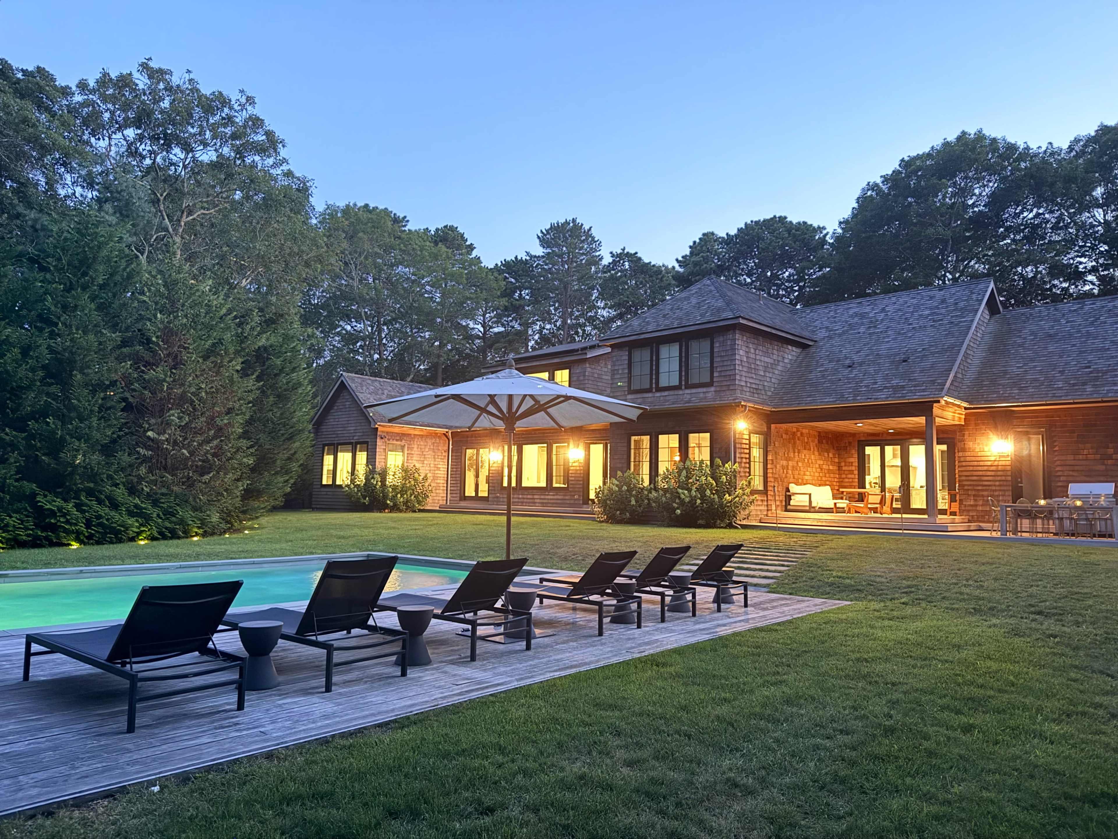The image shows a modern house with a pool and patio area, surrounded by trees in the early evening light.