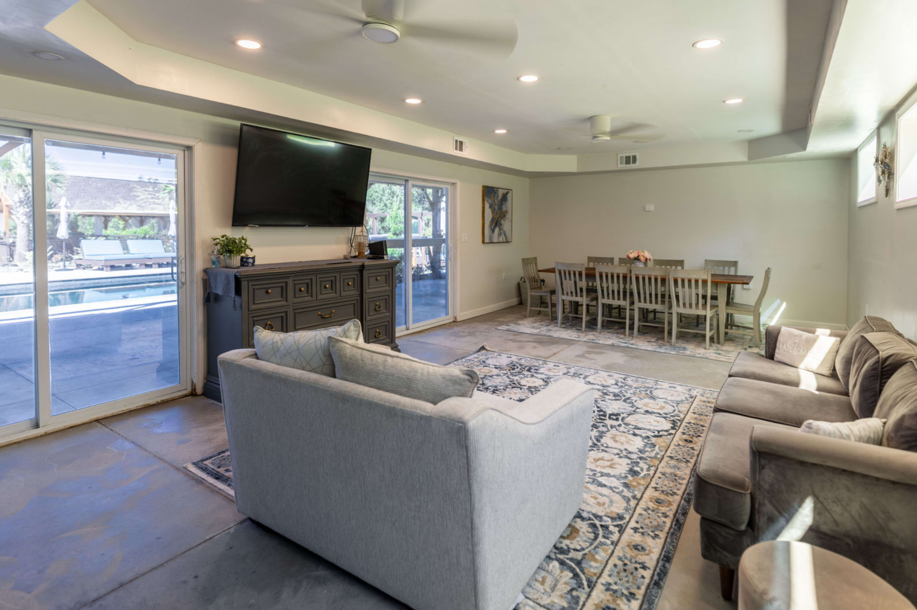 The image shows a spacious living area with a gray couch, a light-colored armchair, a large television mounted on the wall, and a dining table set for several people, with large windows overlooking an outdoor space.