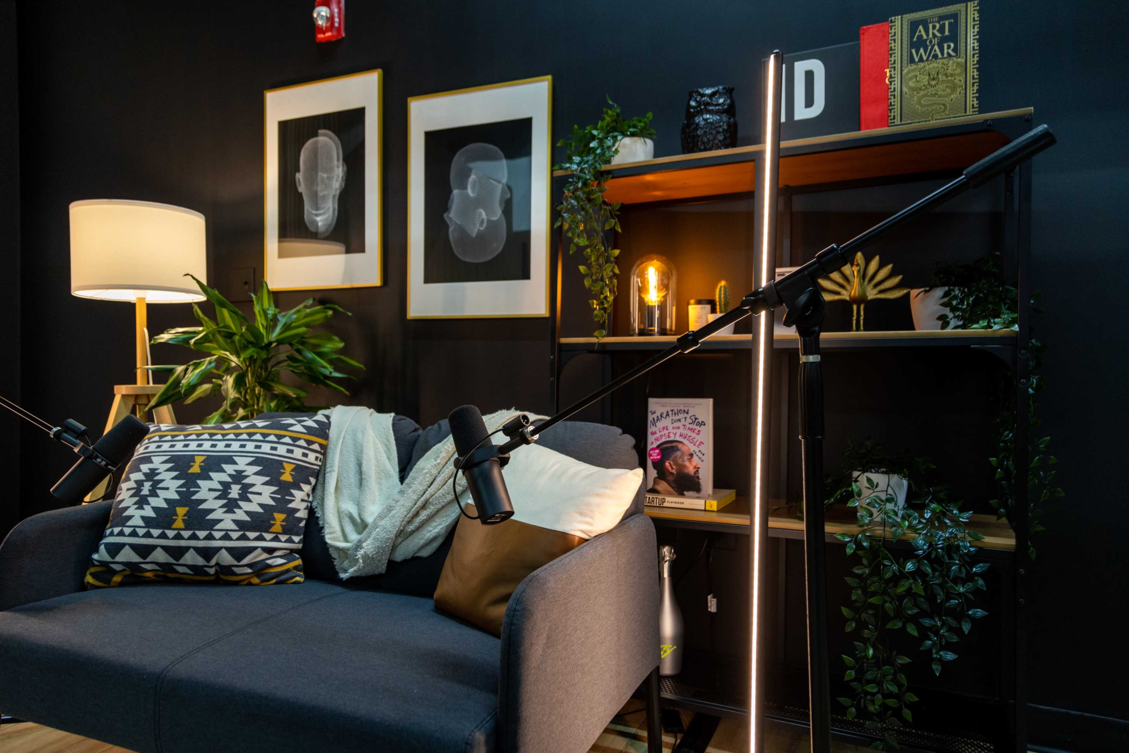 A cozy recording studio setup featuring a gray sofa with throw pillows, two microphones on stands, plants, and shelves displaying books and decor.