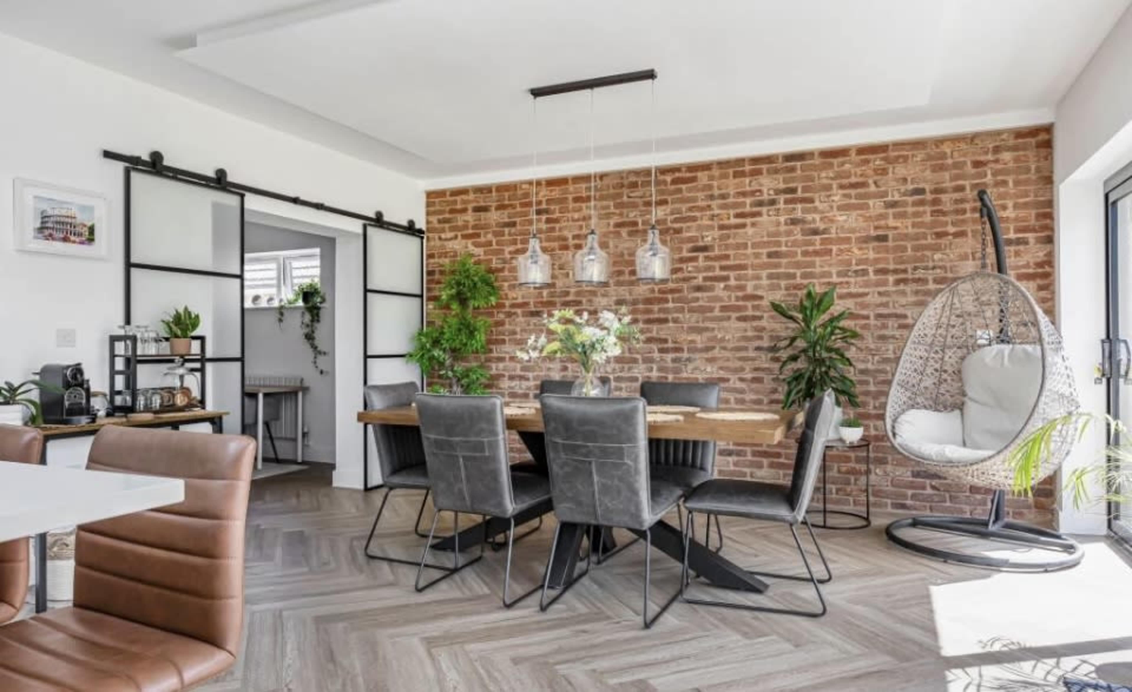 The image shows a dining area with a wooden table surrounded by black chairs, brick walls, and hanging light fixtures, featuring a hanging chair and potted plants.