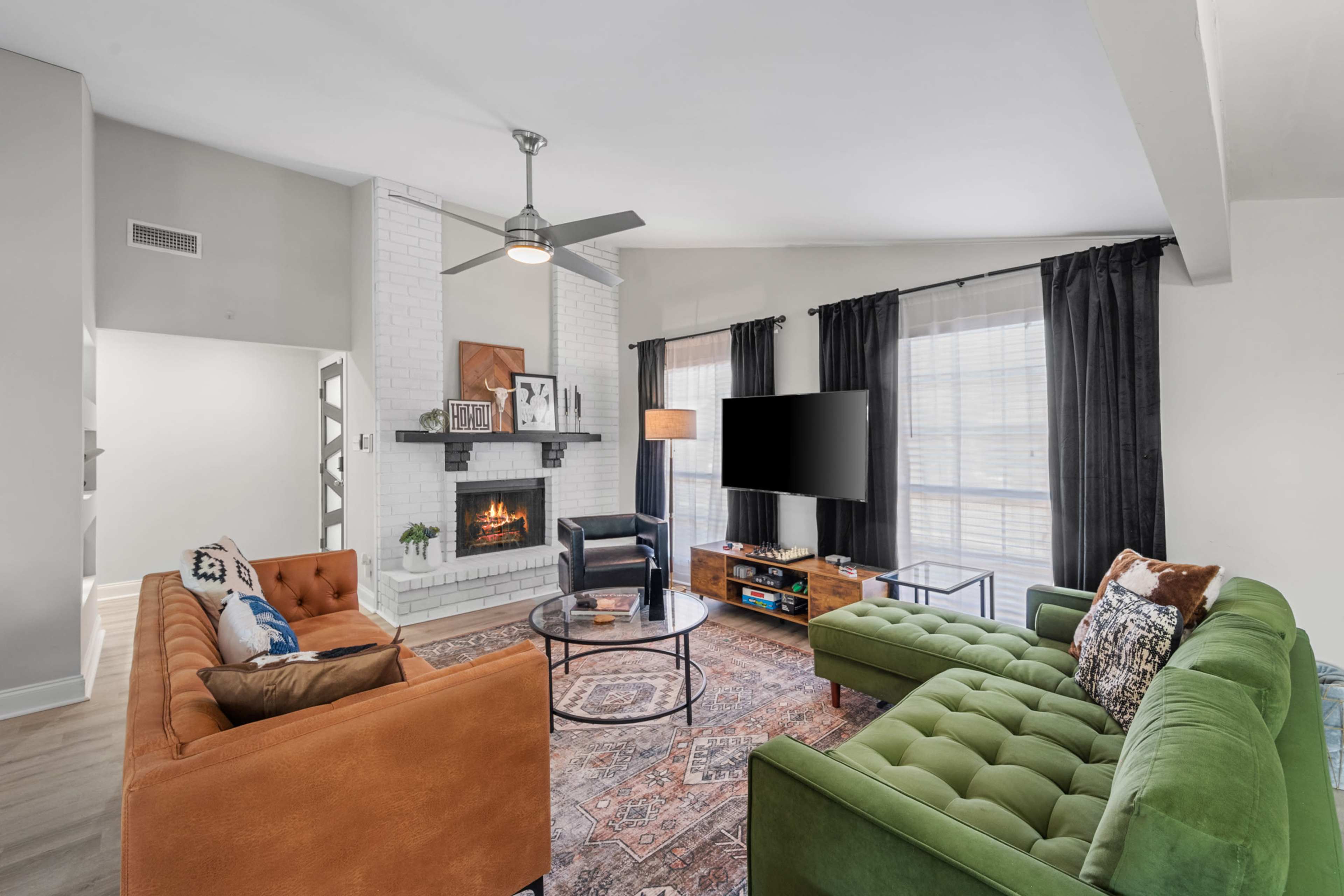 A living room features a cozy seating arrangement with an orange and green couch, a round coffee table, a white brick fireplace, and a television mounted on the wall.