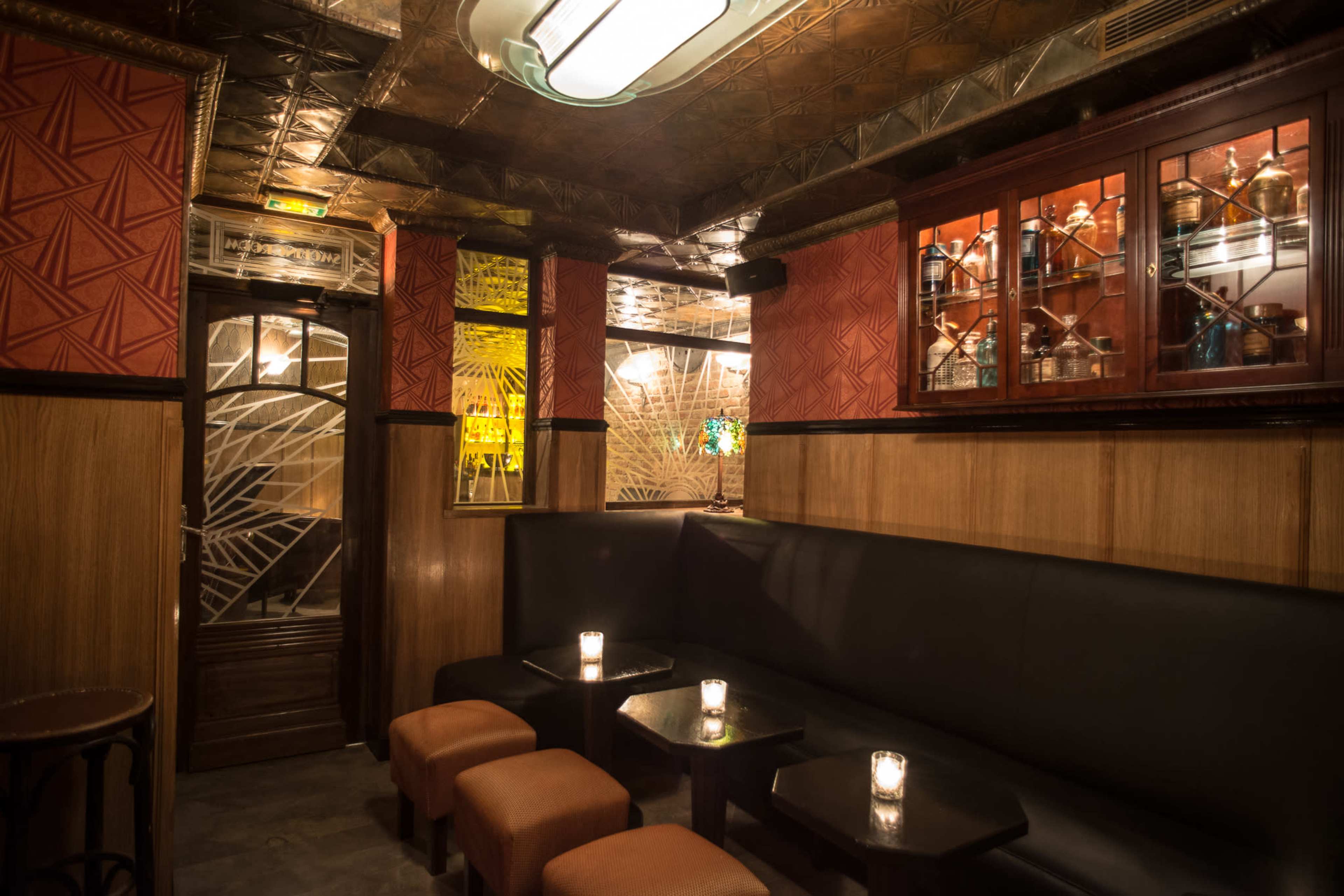 The image shows a cozy bar interior with black seating, wooden accents, glass shelving displaying various items, and dim lighting from small candles on the tables.
