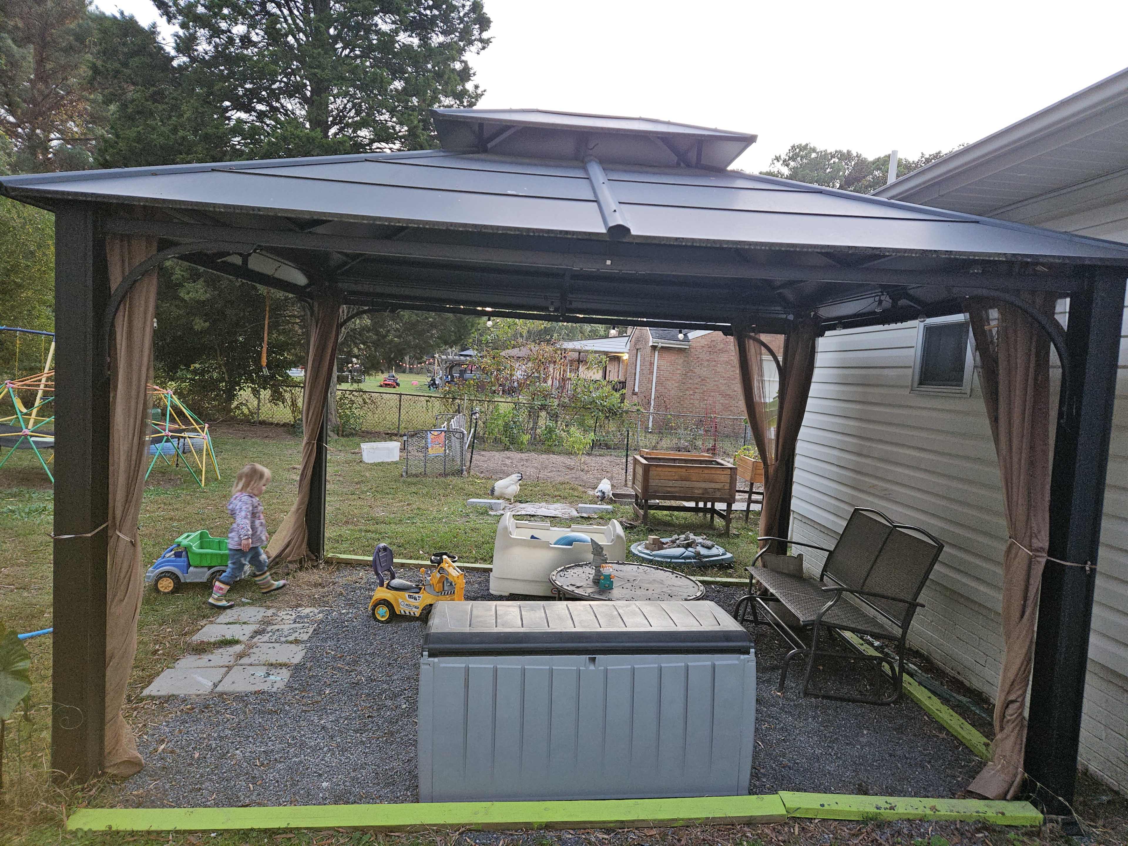 A metal gazebo with mesh curtains stands in a yard, surrounded by children's toys and a grassy area.