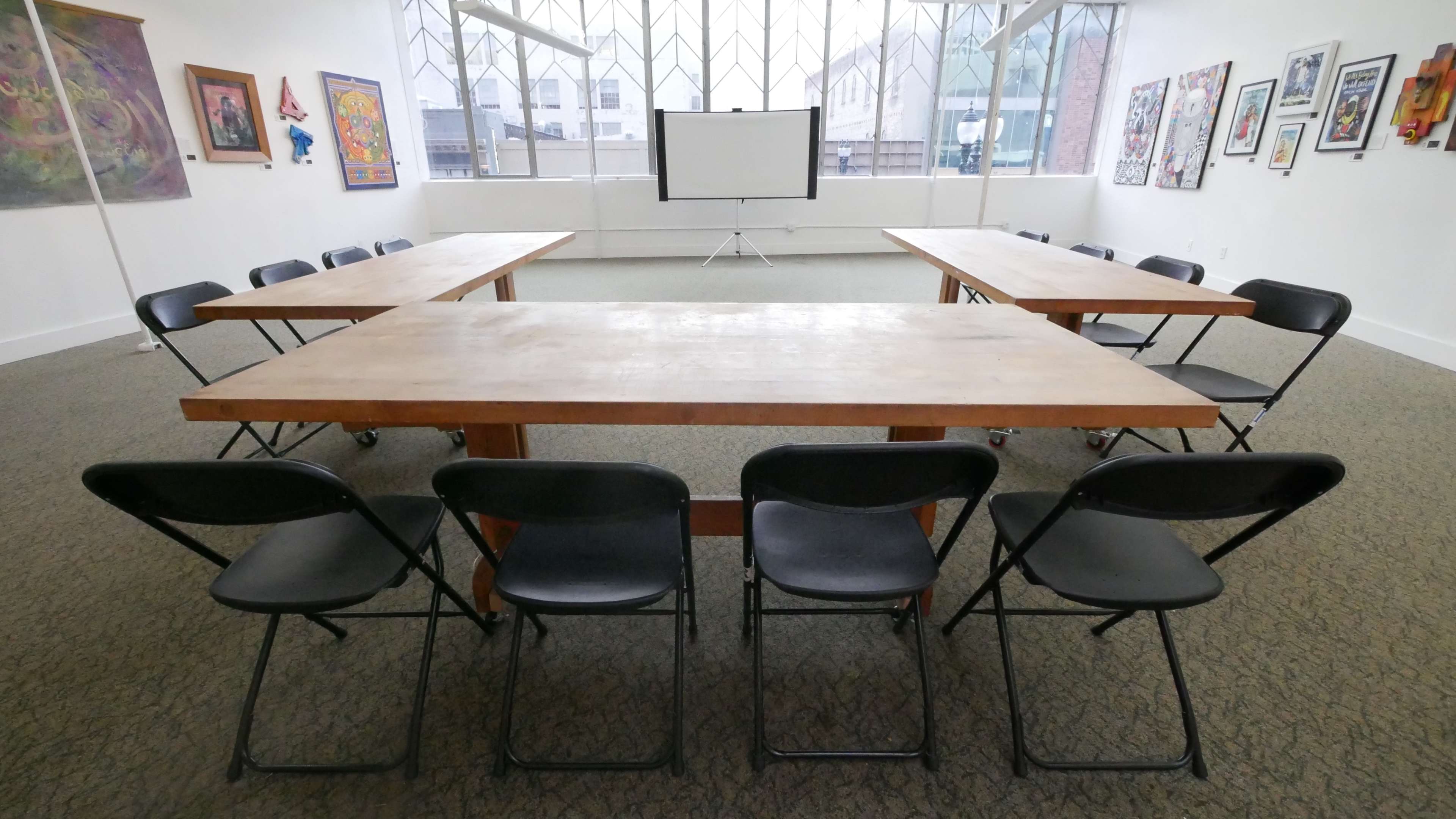 A spacious room features two large, rectangular tables arranged in a U-shape, surrounded by black folding chairs, with a projector screen positioned at the far end.