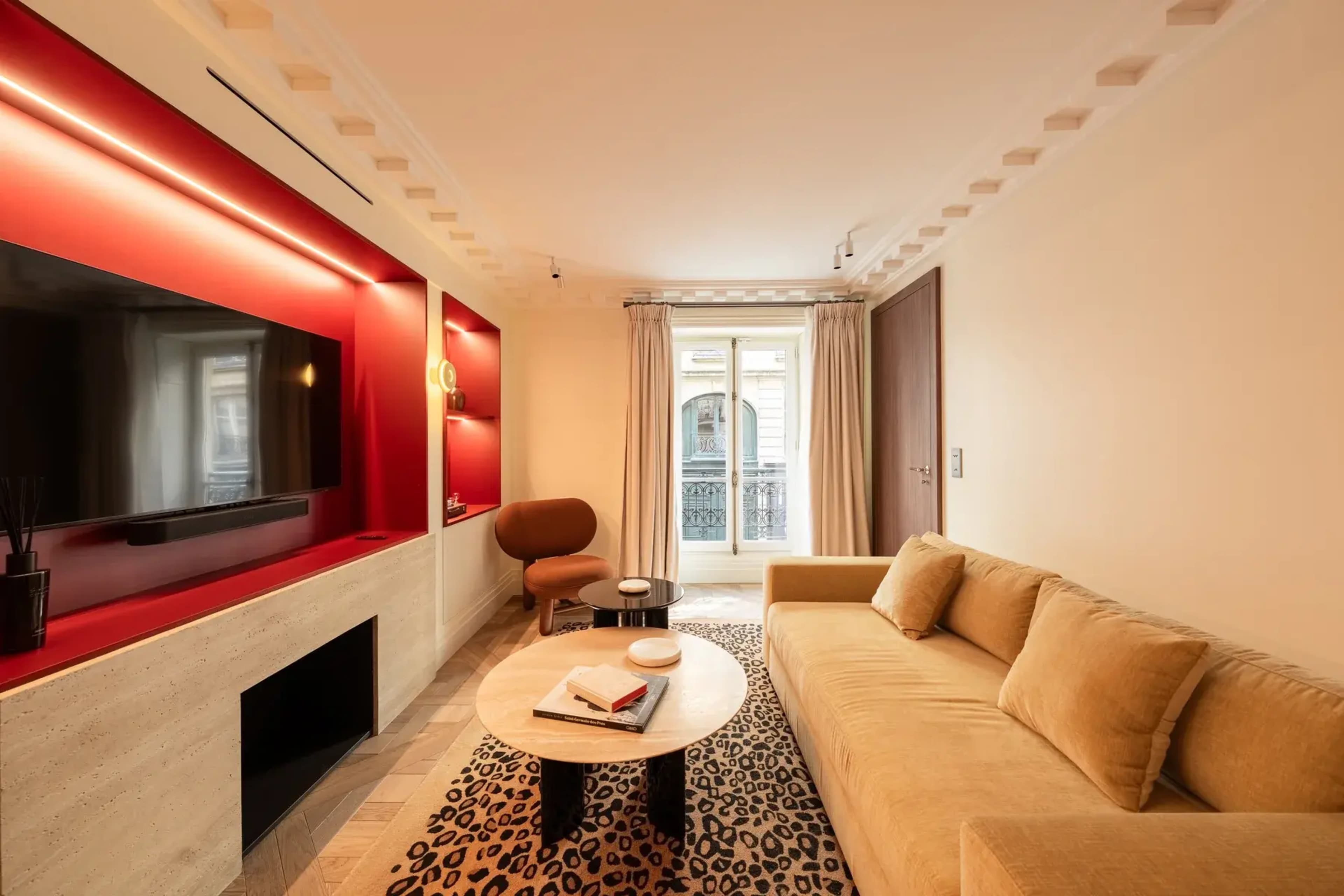 The image depicts a living room with a beige sofa, a round coffee table on a patterned rug, and a wall-mounted television set against a feature wall with red accents.