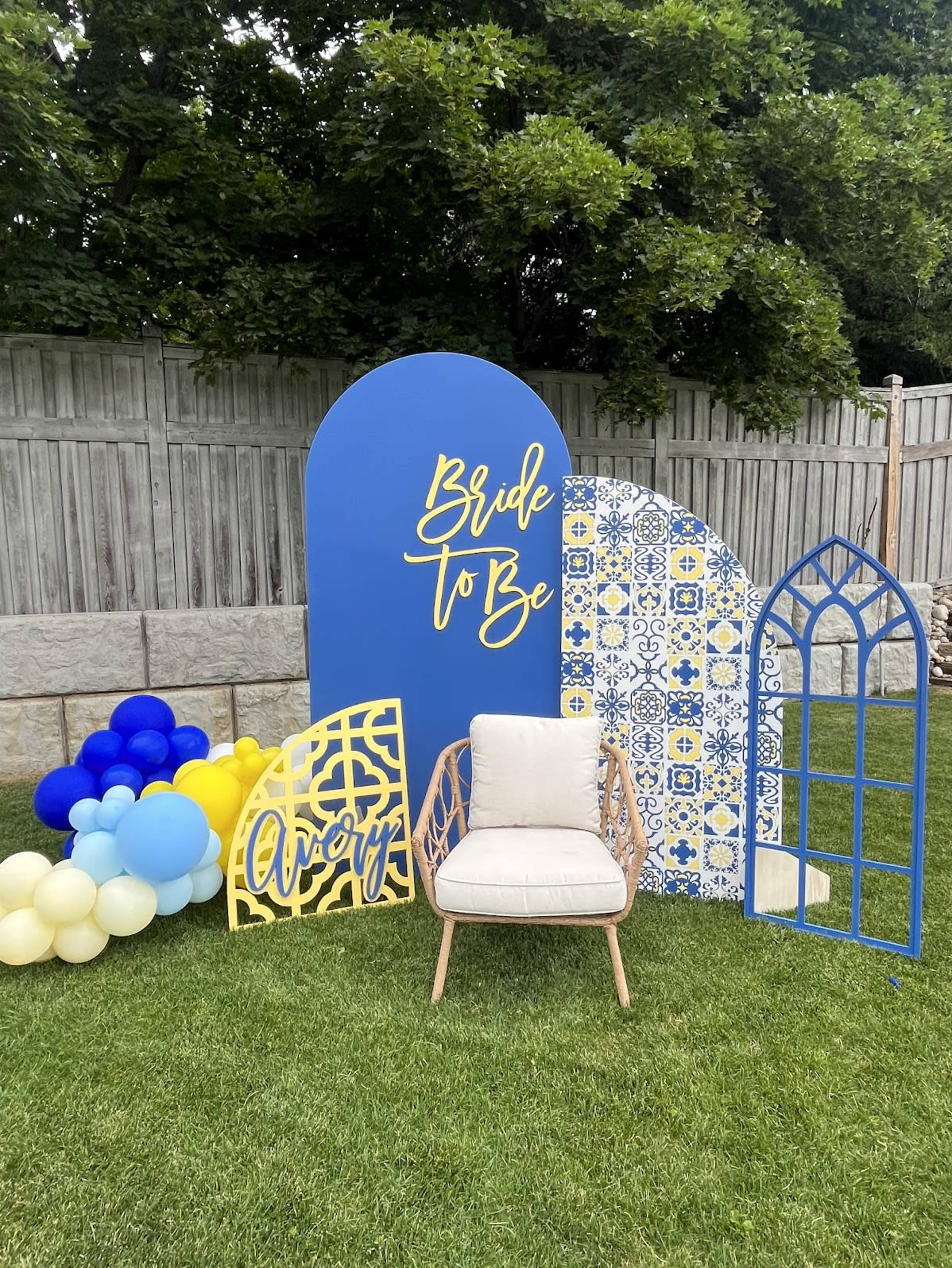 The image shows a decorated outdoor area featuring a "Bride To Be" sign, colorful arch backdrops, and a chair surrounded by balloons on a grassy surface.