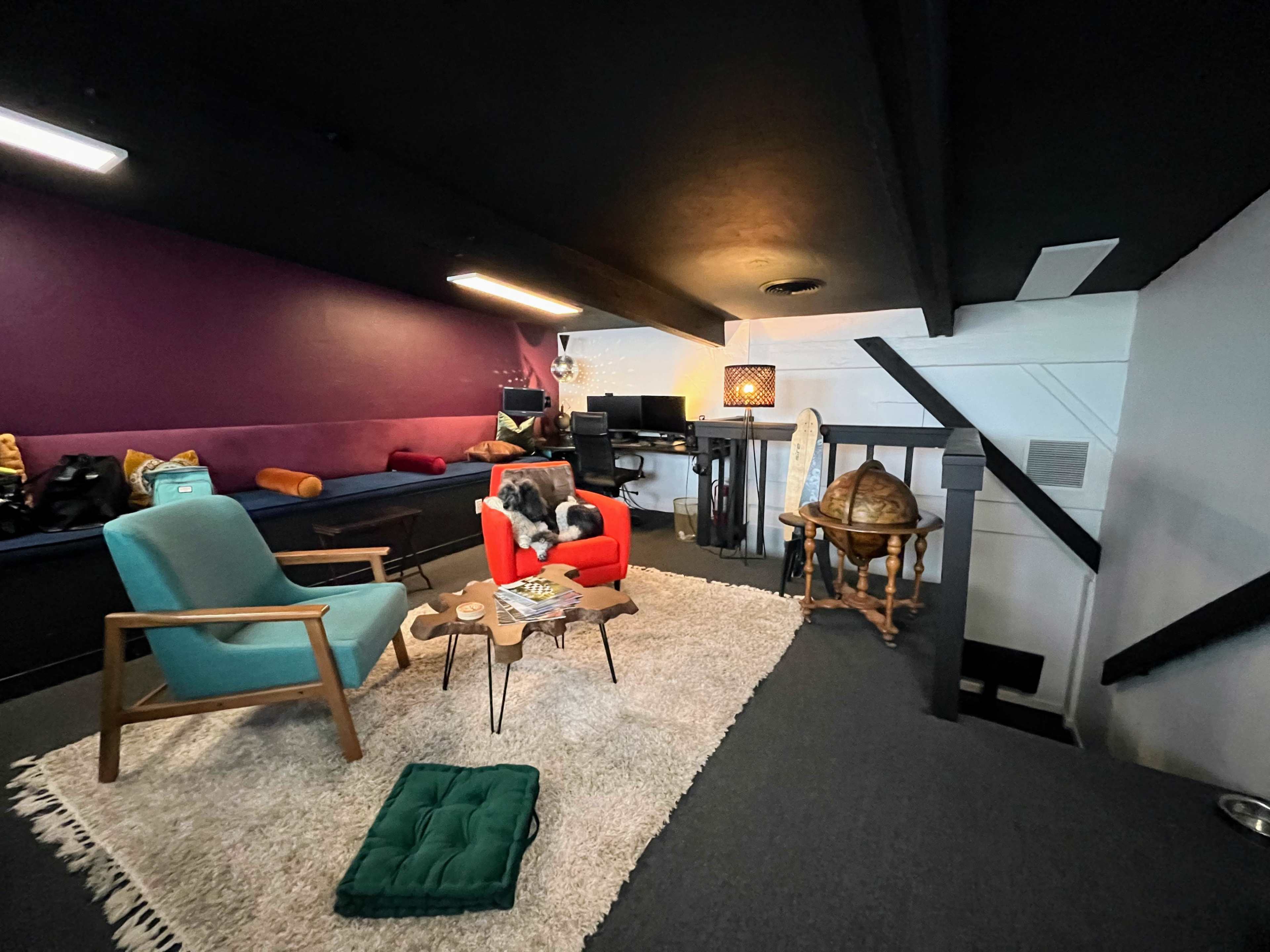 The image shows a cozy interior space featuring a red armchair, a green mat, a coffee table with decor items, and various seating arrangements against a purple and white wall backdrop.