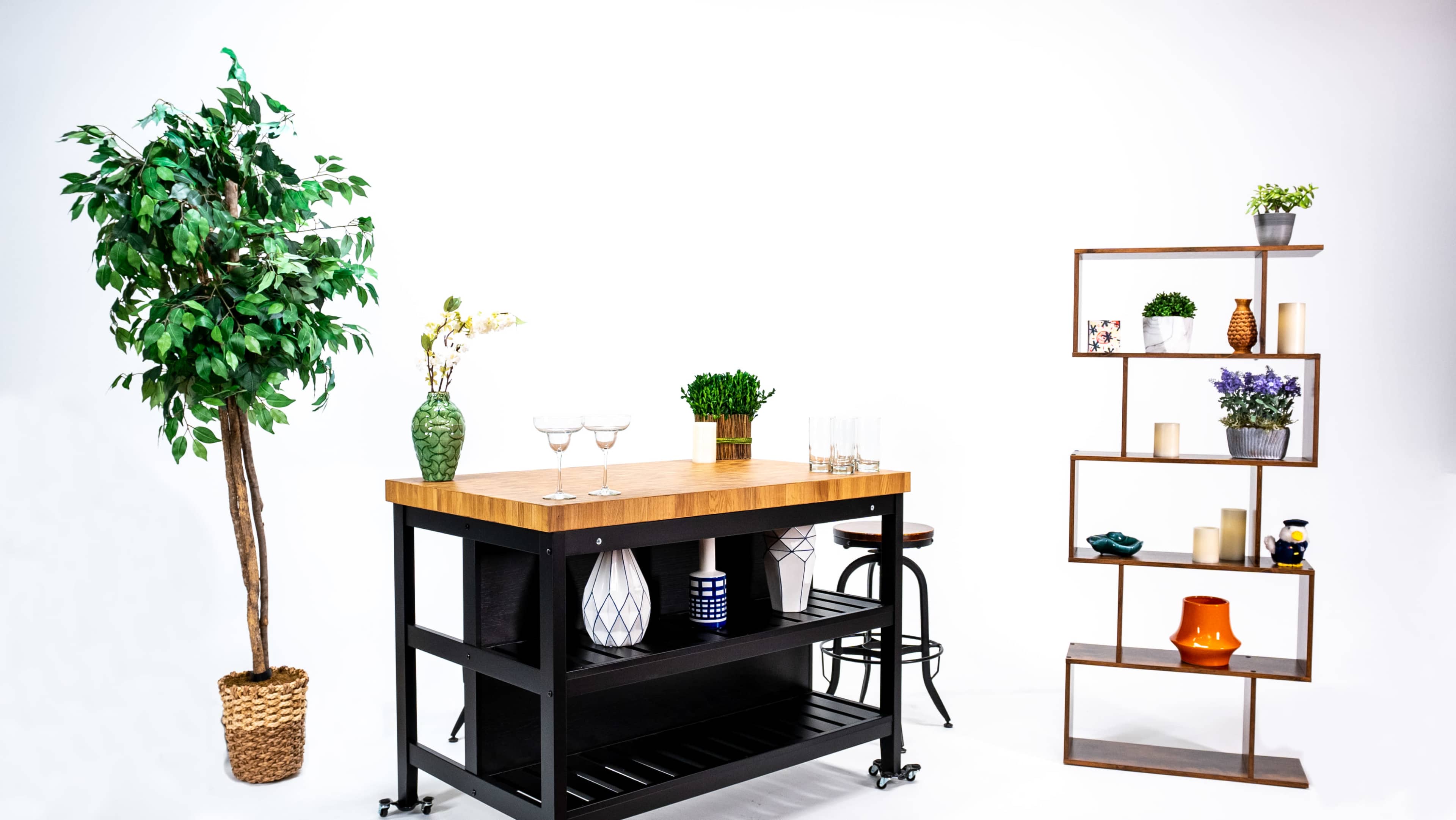 A small bar cart with a wooden top and various decorative items, accompanied by a potted plant and a modern shelving unit against a white background.
