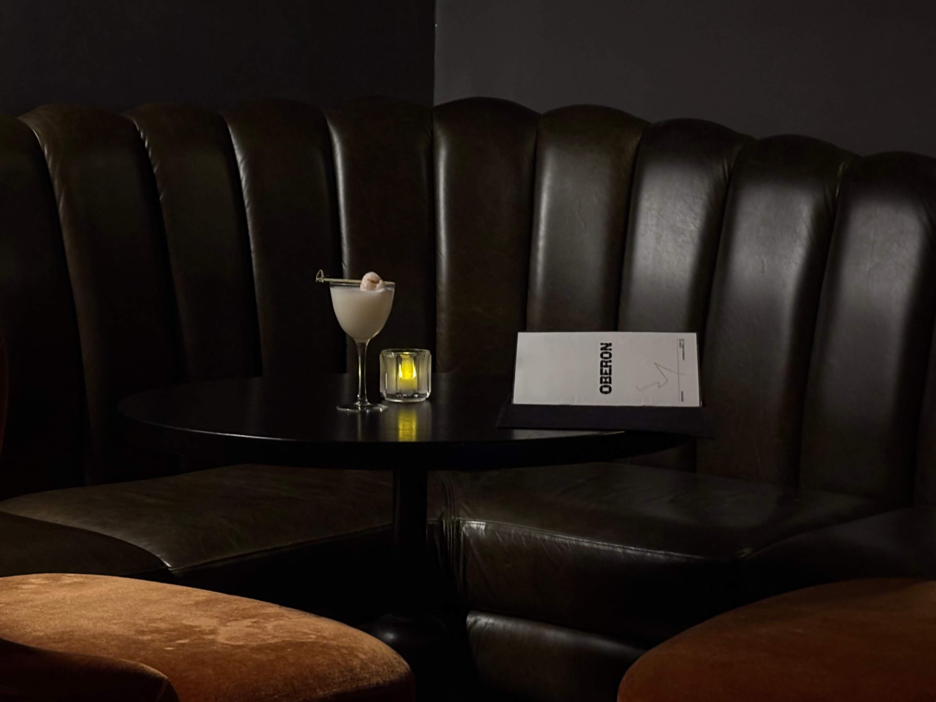 A small, dimly lit table in a cozy seating area features a cocktail and a menu beside a glowing candle.