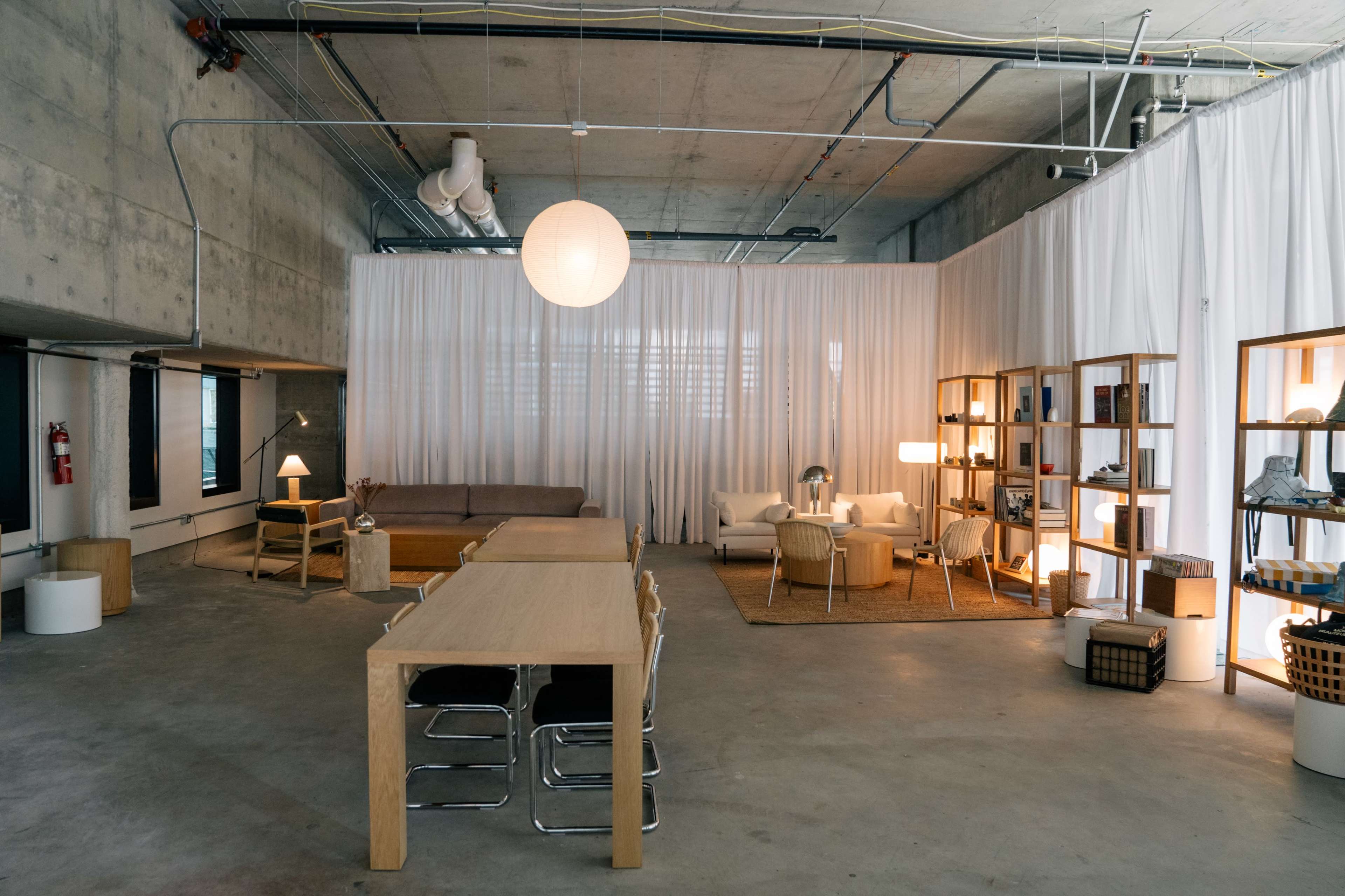 The image shows a modern, open-concept space with a large table, chairs, a sofa, and bookshelves, separated by sheer curtains.