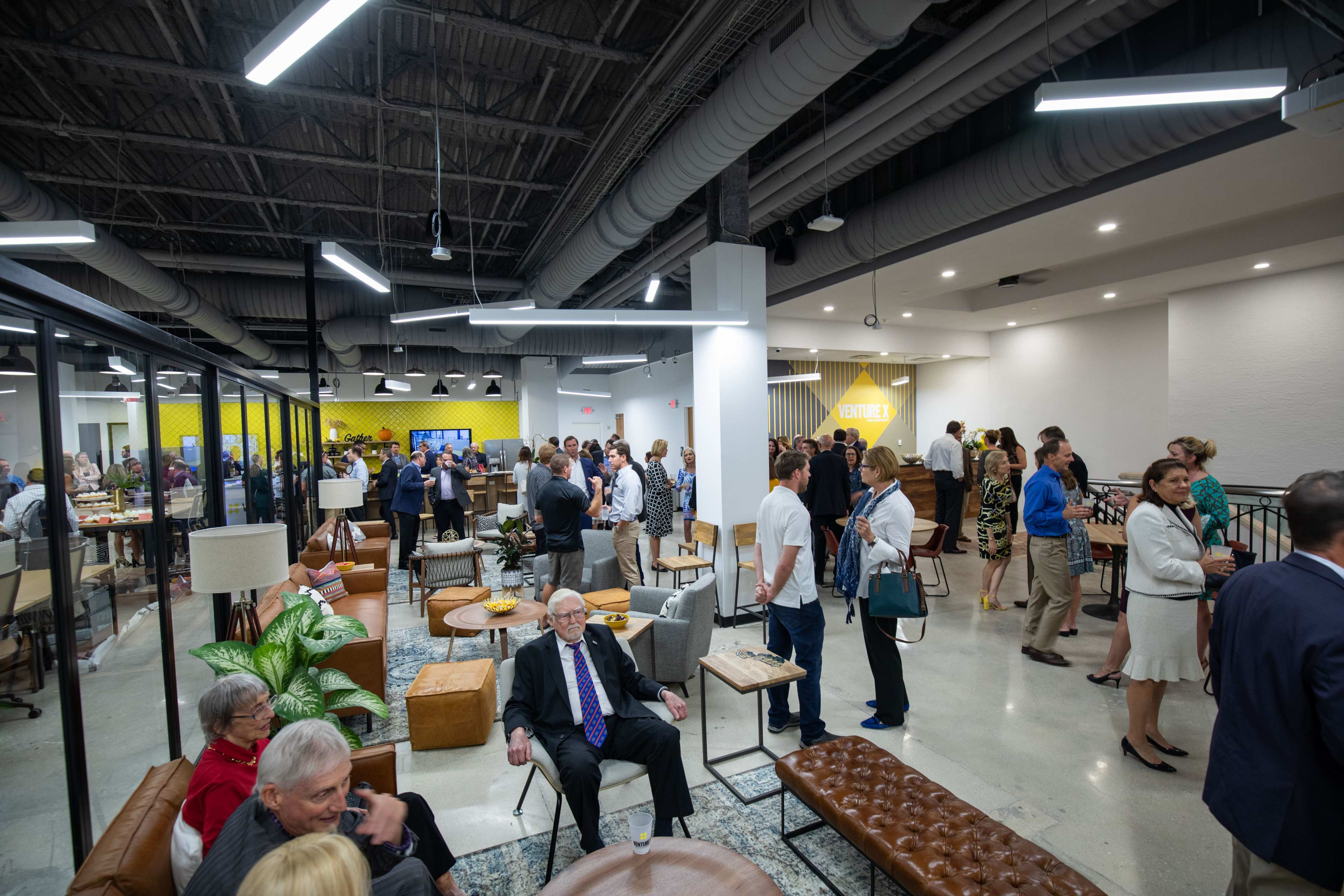 A large group of people gathers in a modern office space filled with seating areas and bright decor, engaging in conversation and networking.