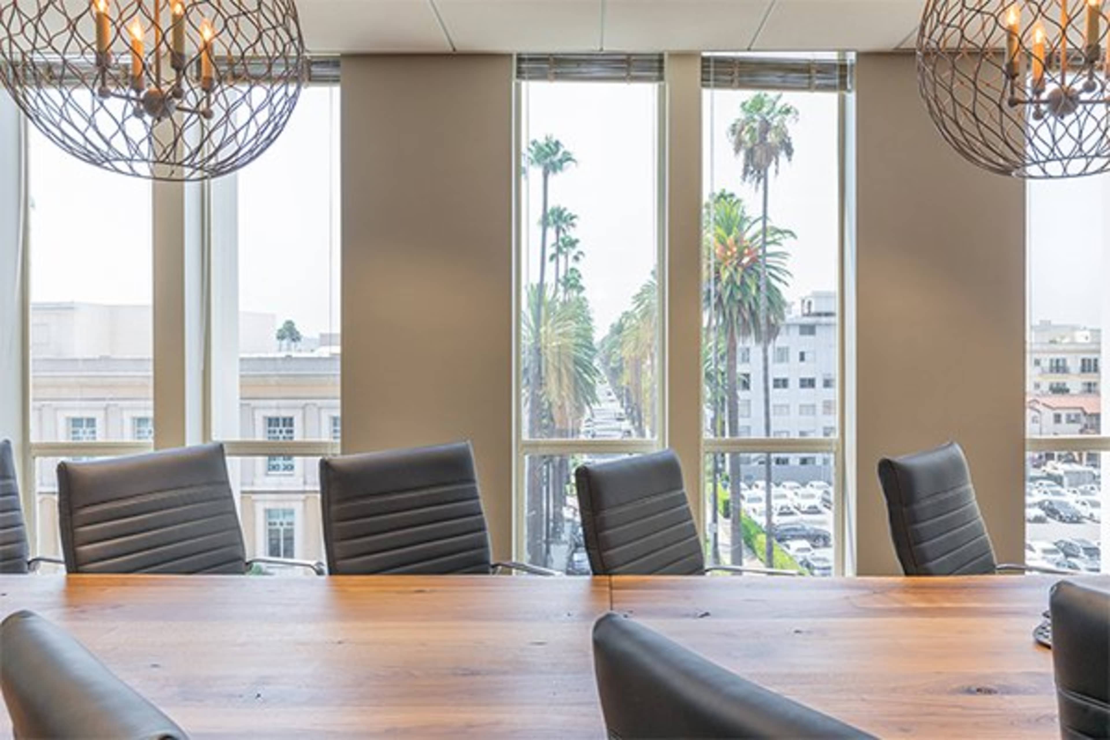 A conference room features a large wooden table surrounded by black chairs, with large windows displaying palm trees and a street outside.