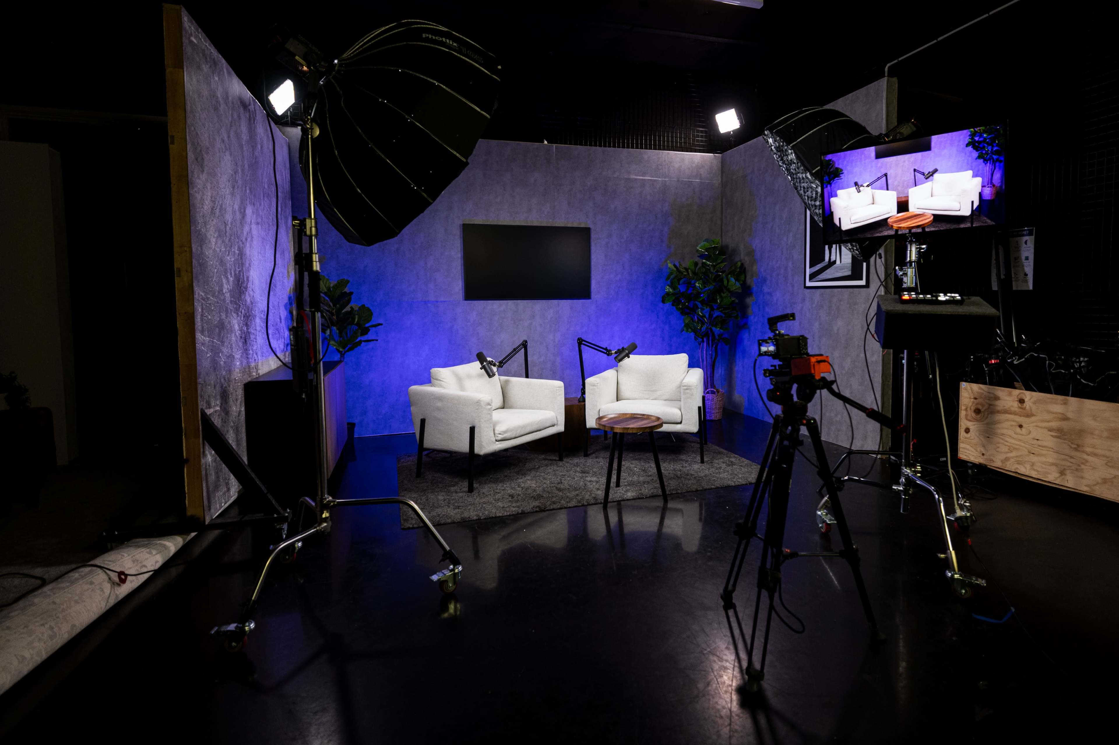 A modern studio setup features two white chairs, a small wooden table, and lighting equipment against a backdrop of neutral colors and decorative plants.