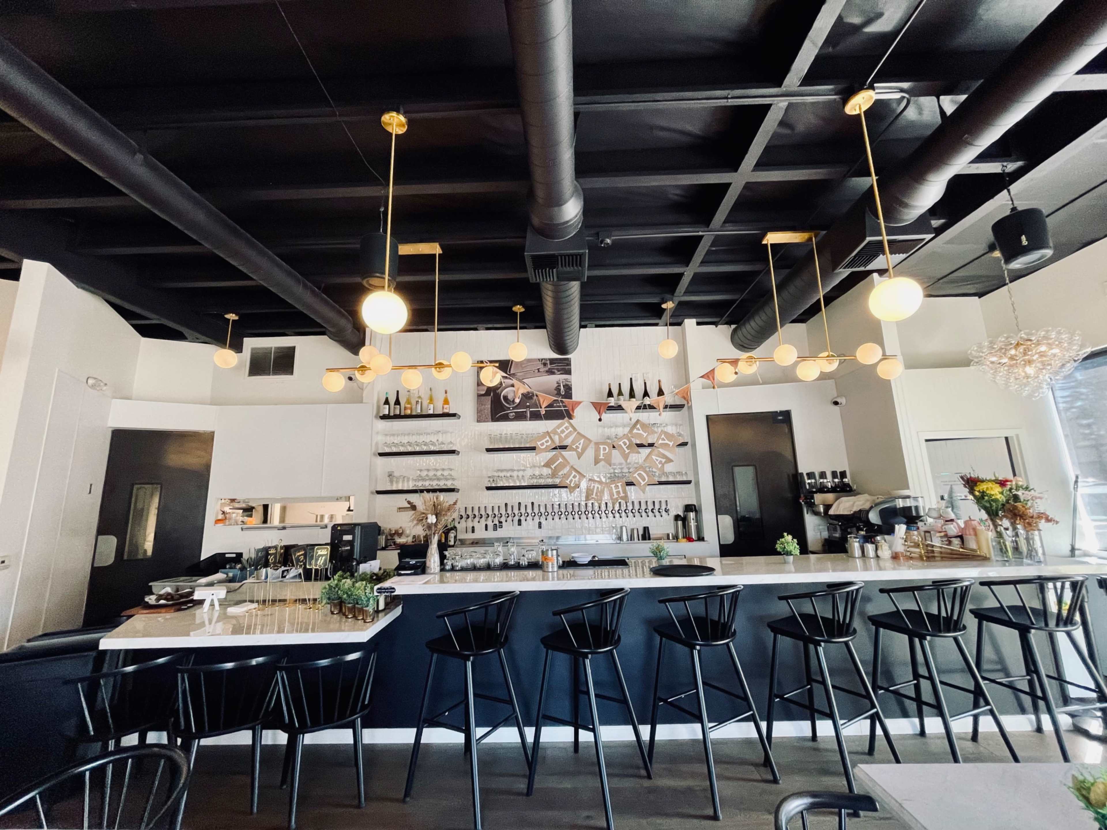The image shows a modern bar area with a sleek counter, high black stools, and shelves displaying various bottles and decorations.