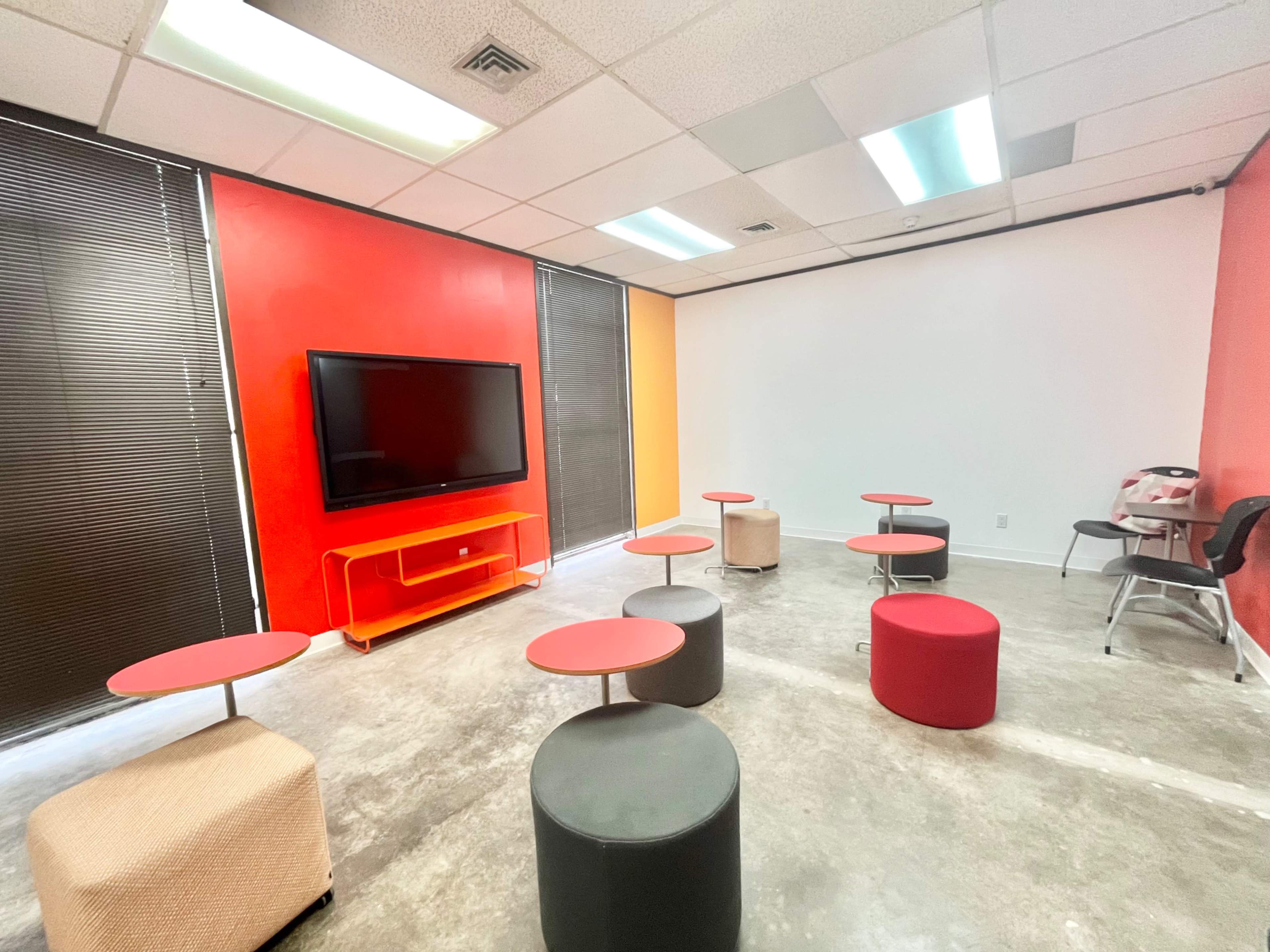 The image shows a room with a TV mounted on the wall and several seating arrangements consisting of small tables and stools in a mix of red and gray colors.