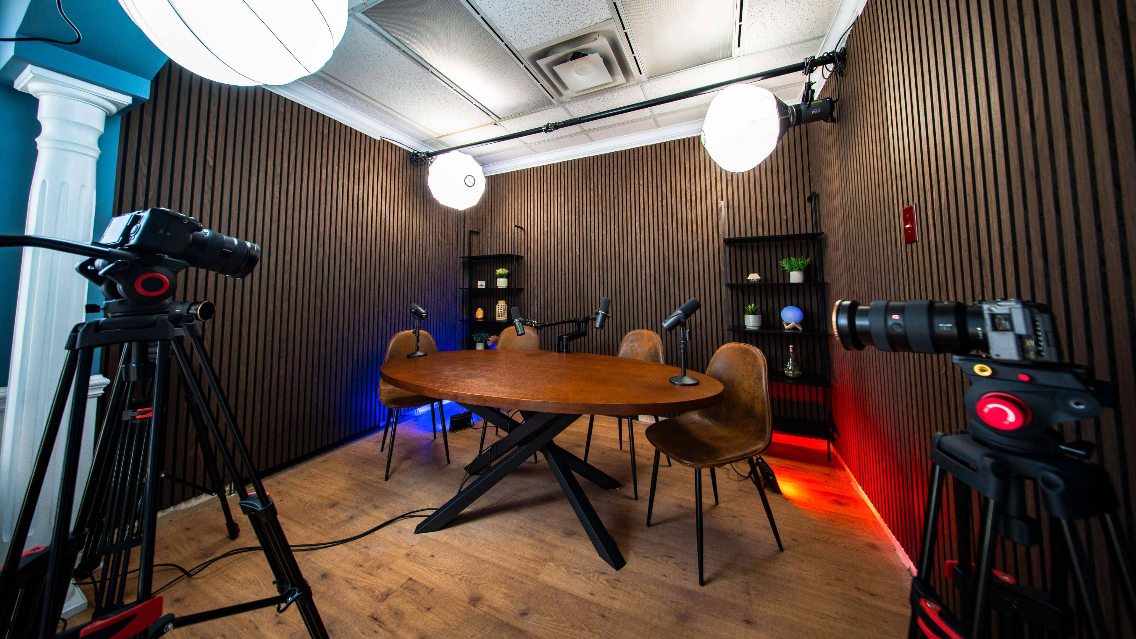 The image shows a recording studio with a wooden table and chairs, surrounded by cameras and lighting equipment against a background of wooden paneling.